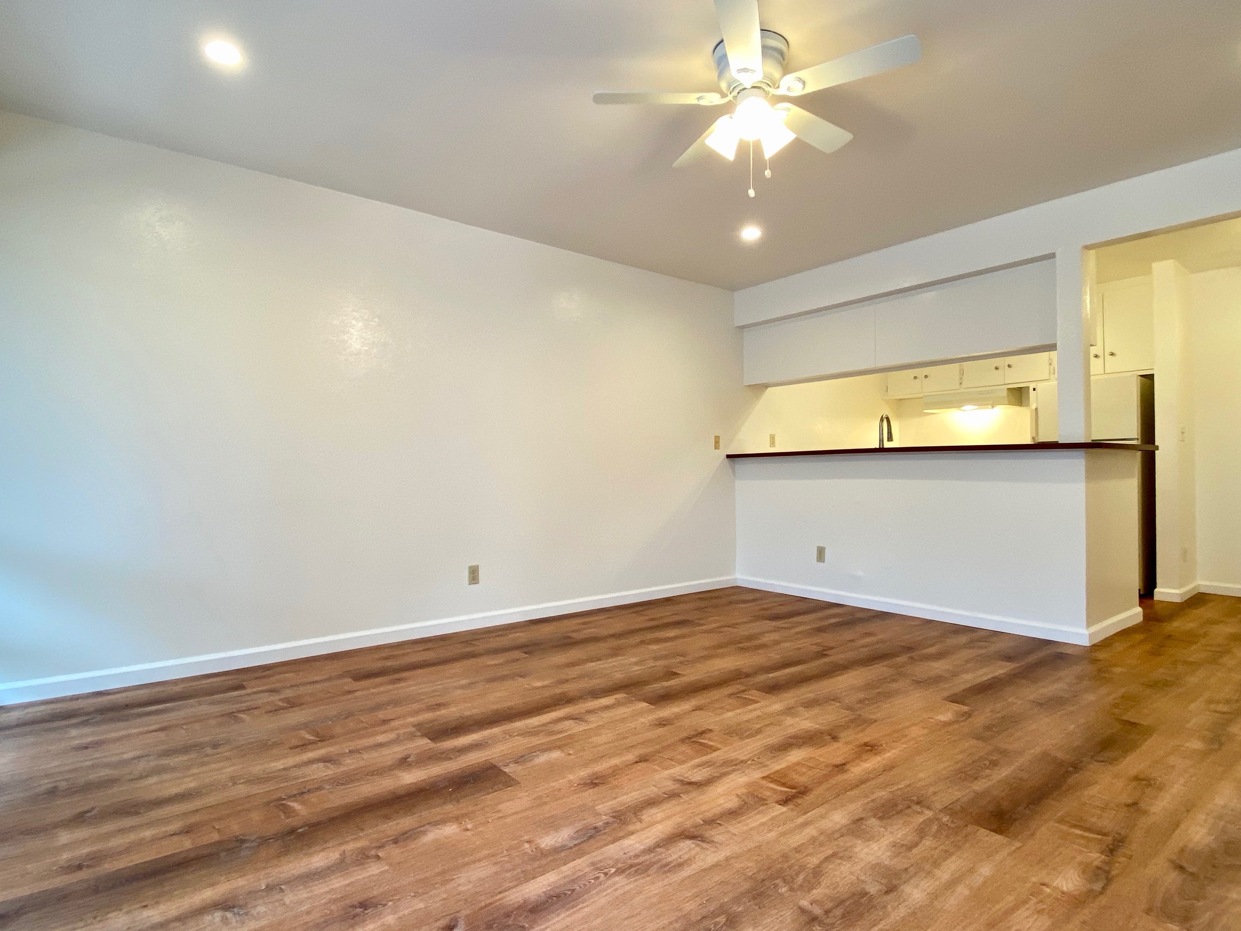 Empty living room with white walls, ceiling fan, and hardwood flooring.