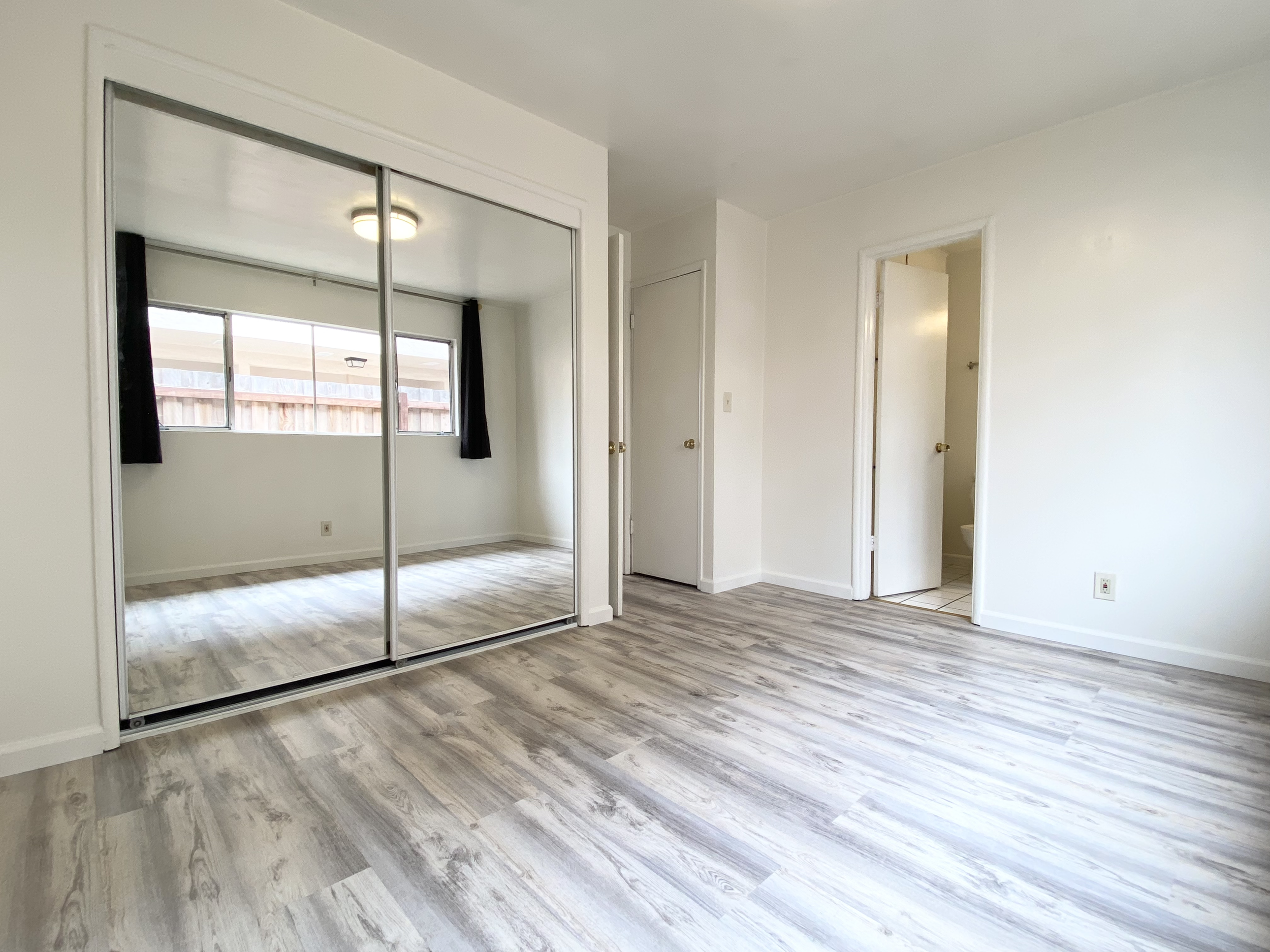 Empty room with wood-like flooring, white walls, closet with mirrored sliding doors, and a window with black curtains.