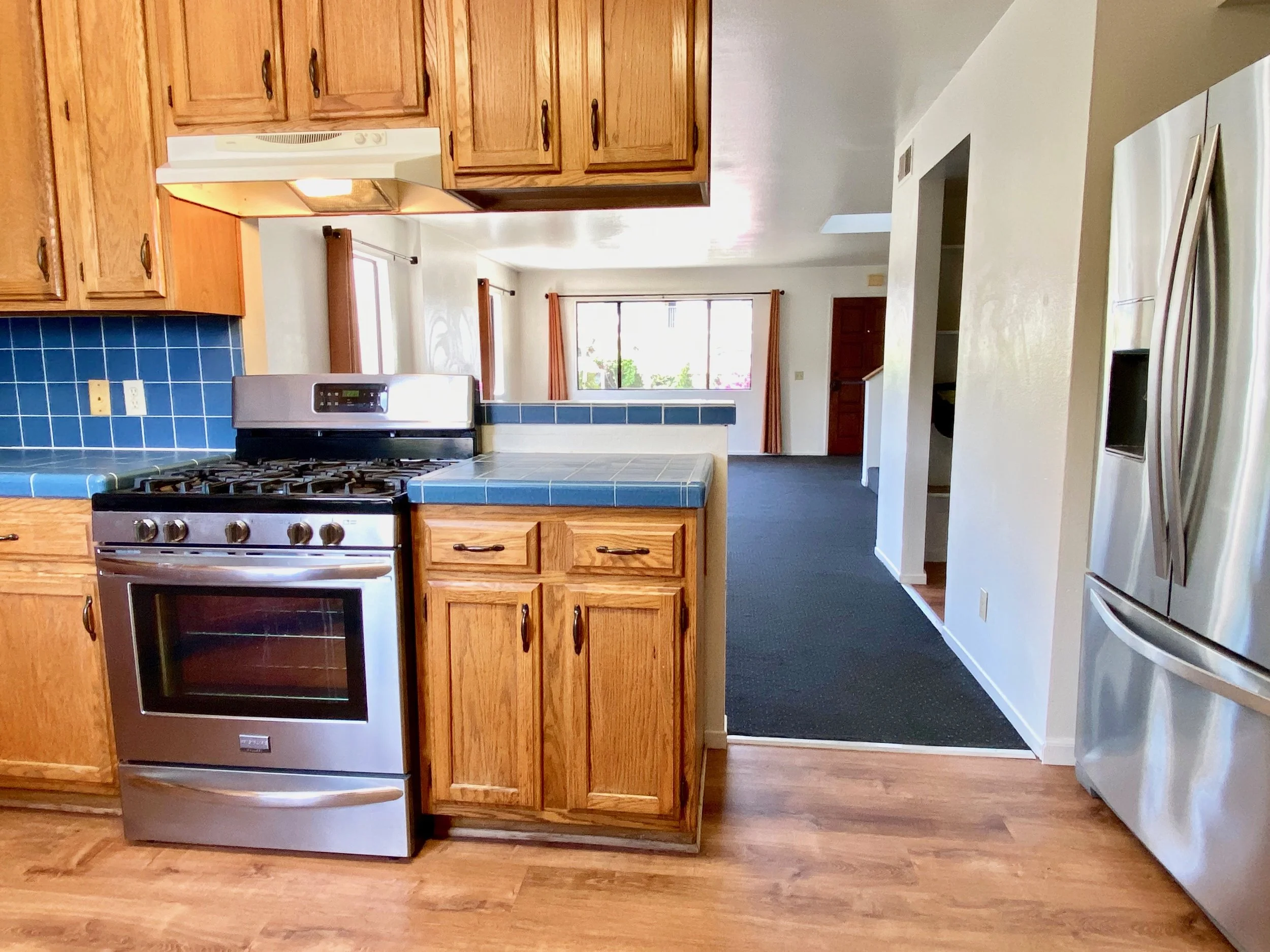 Kitchen with wooden cabinets, blue tiled countertops, a stainless steel stove, and a refrigerator, with a view into a living room with large windows.