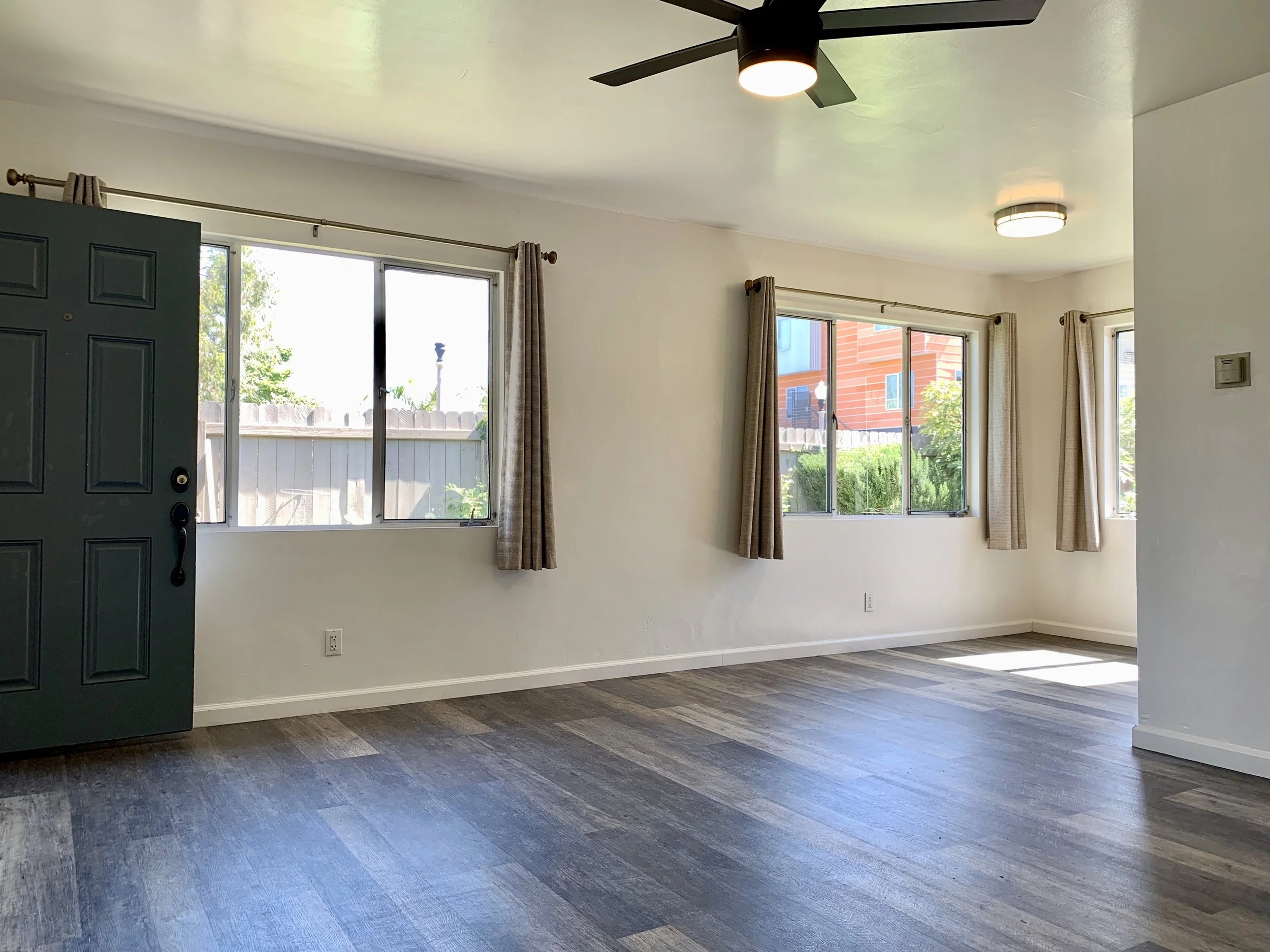 Living room with hardwood floors, large windows with beige curtains, white walls, and a ceiling fan with a light.