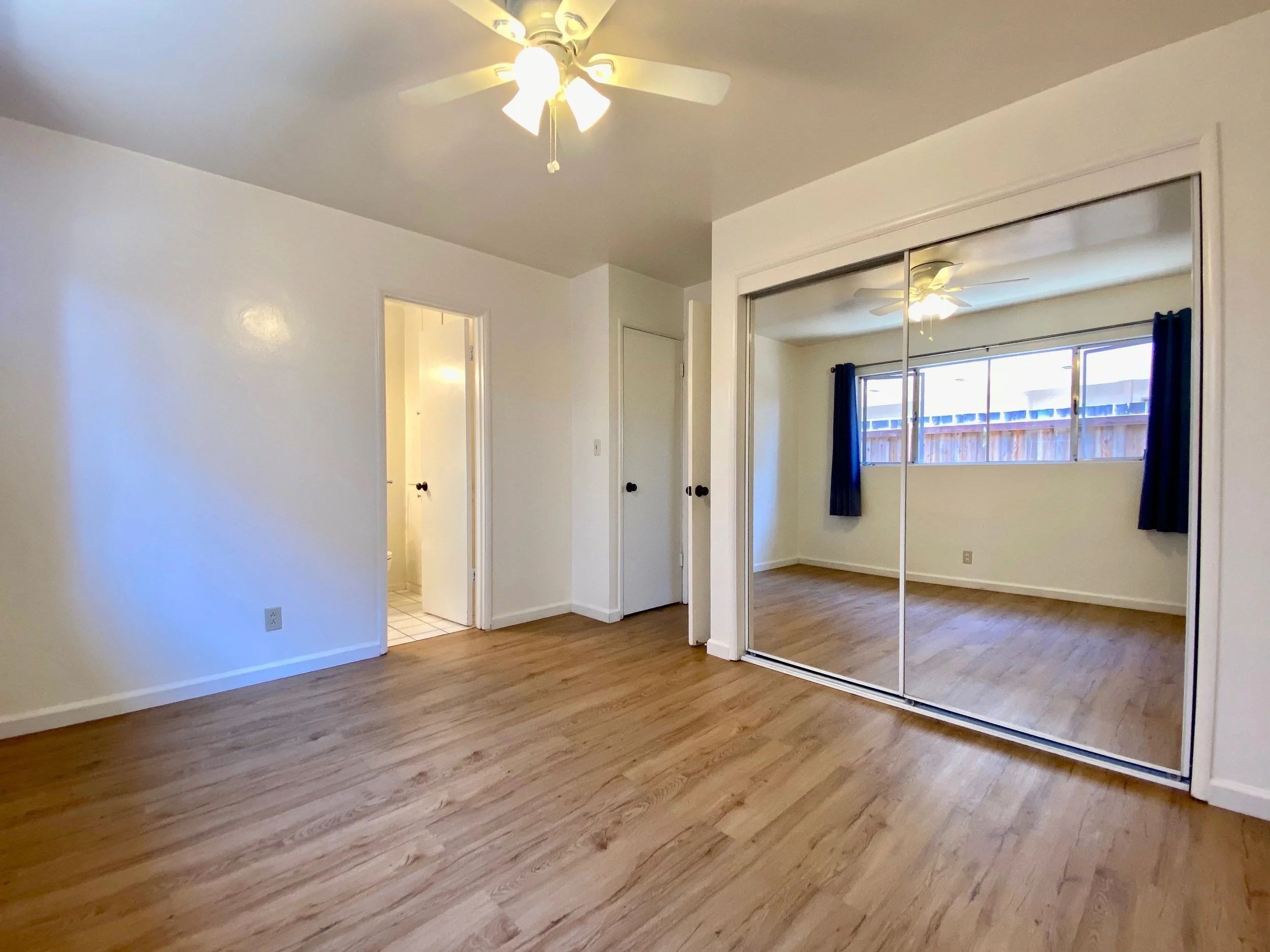 A bedroom with wood flooring, a ceiling fan with lights, mirrored closet doors, a window with blue curtains, and an open door leading to a bathroom.