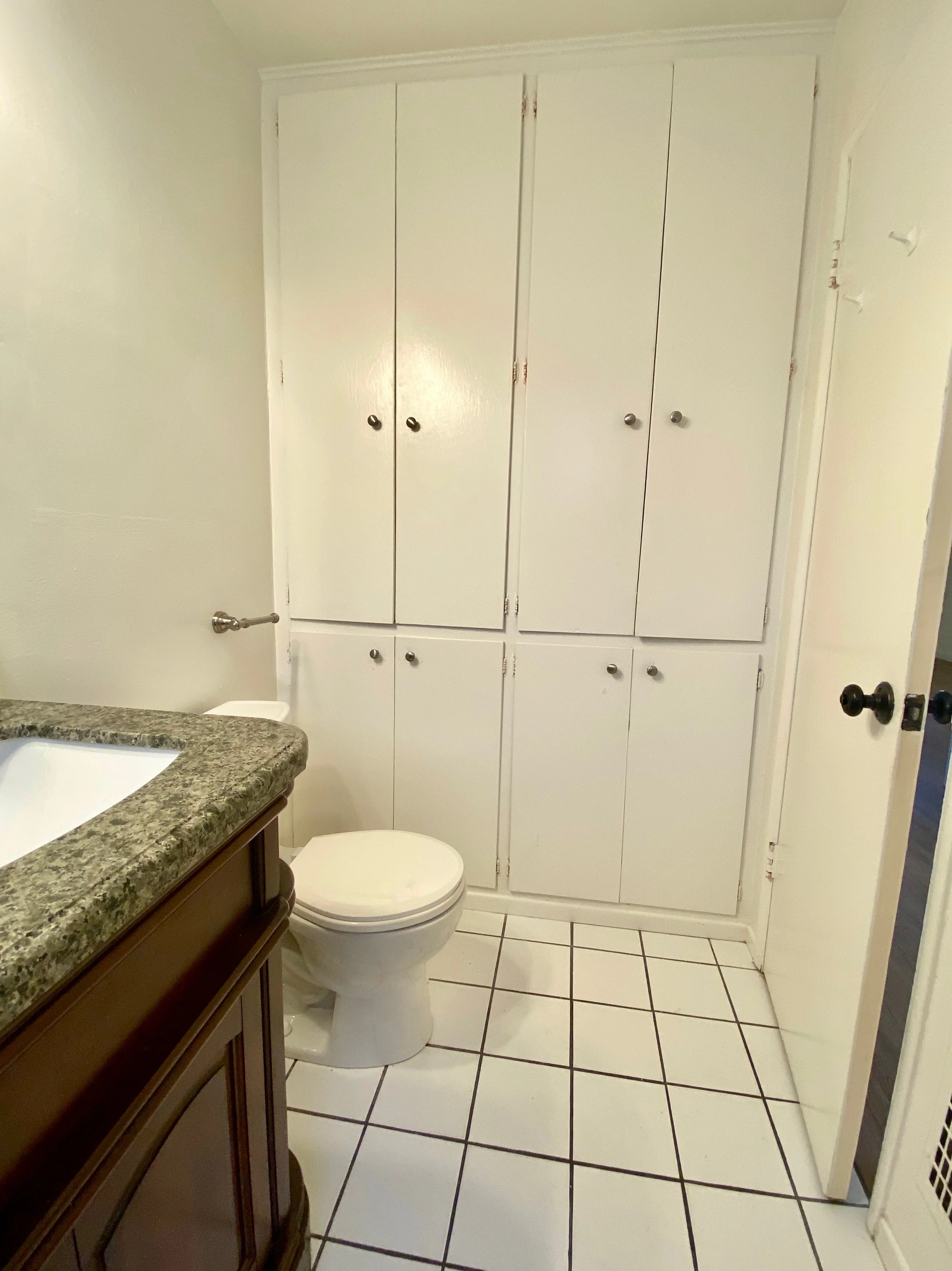 A bathroom with white cabinets, a granite countertop, a toilet, and white tiled floor.