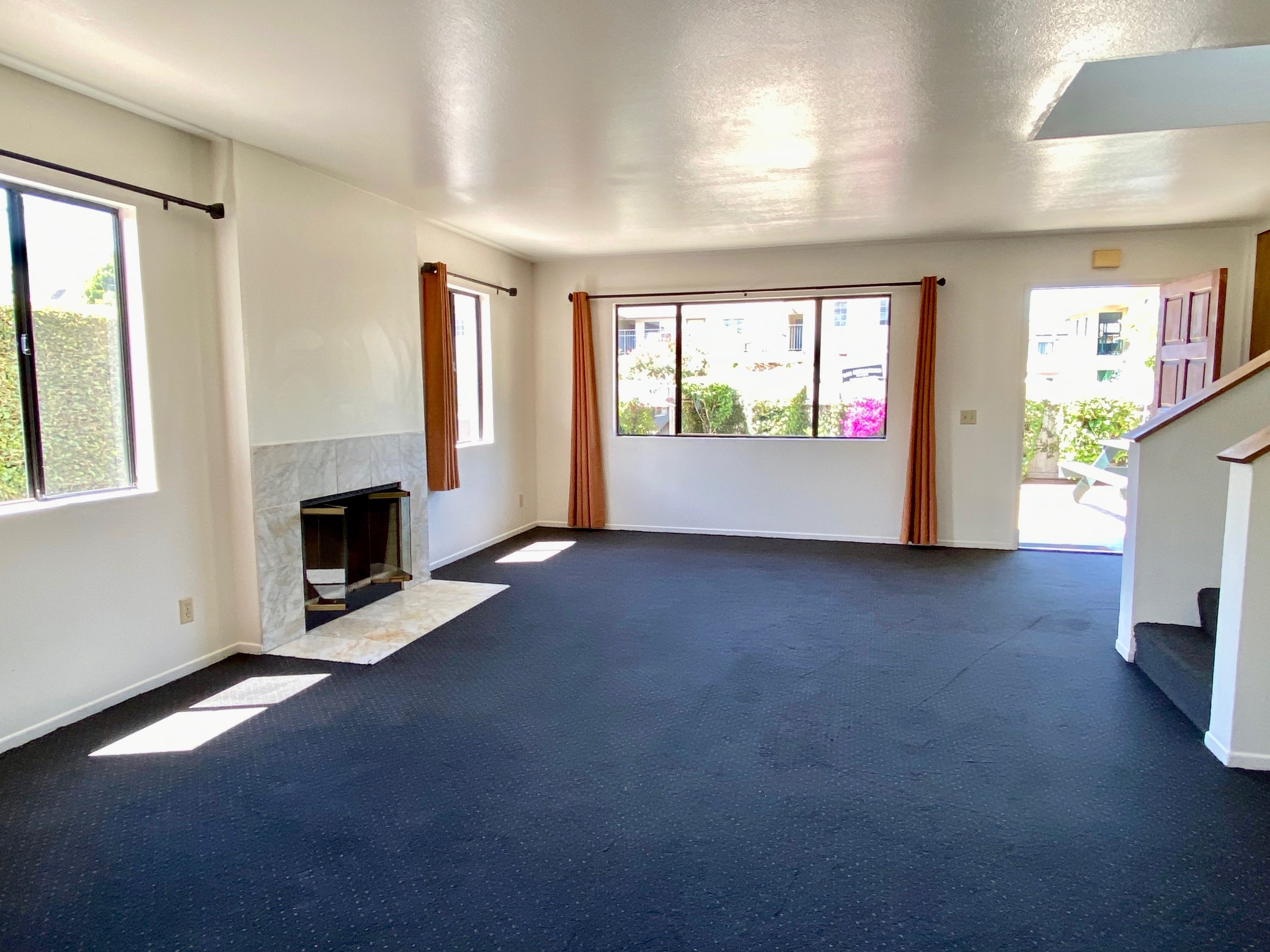 Bright living room with large windows, navy blue carpet, and white walls, featuring a corner fireplace with marble surround and orange curtains.