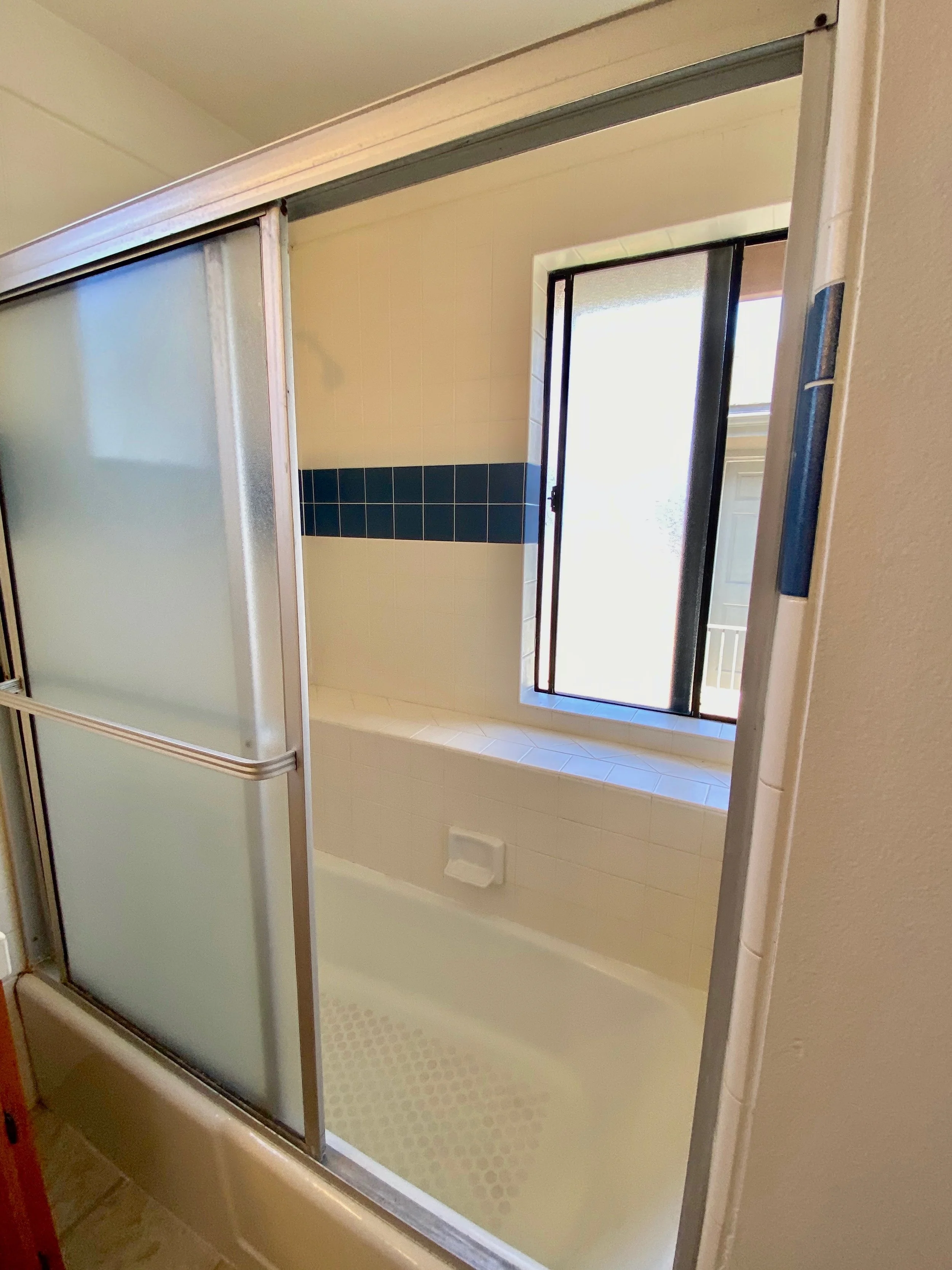 Bathroom shower with sliding frosted glass door, white tile walls with a blue tile accent, and a window providing natural light.