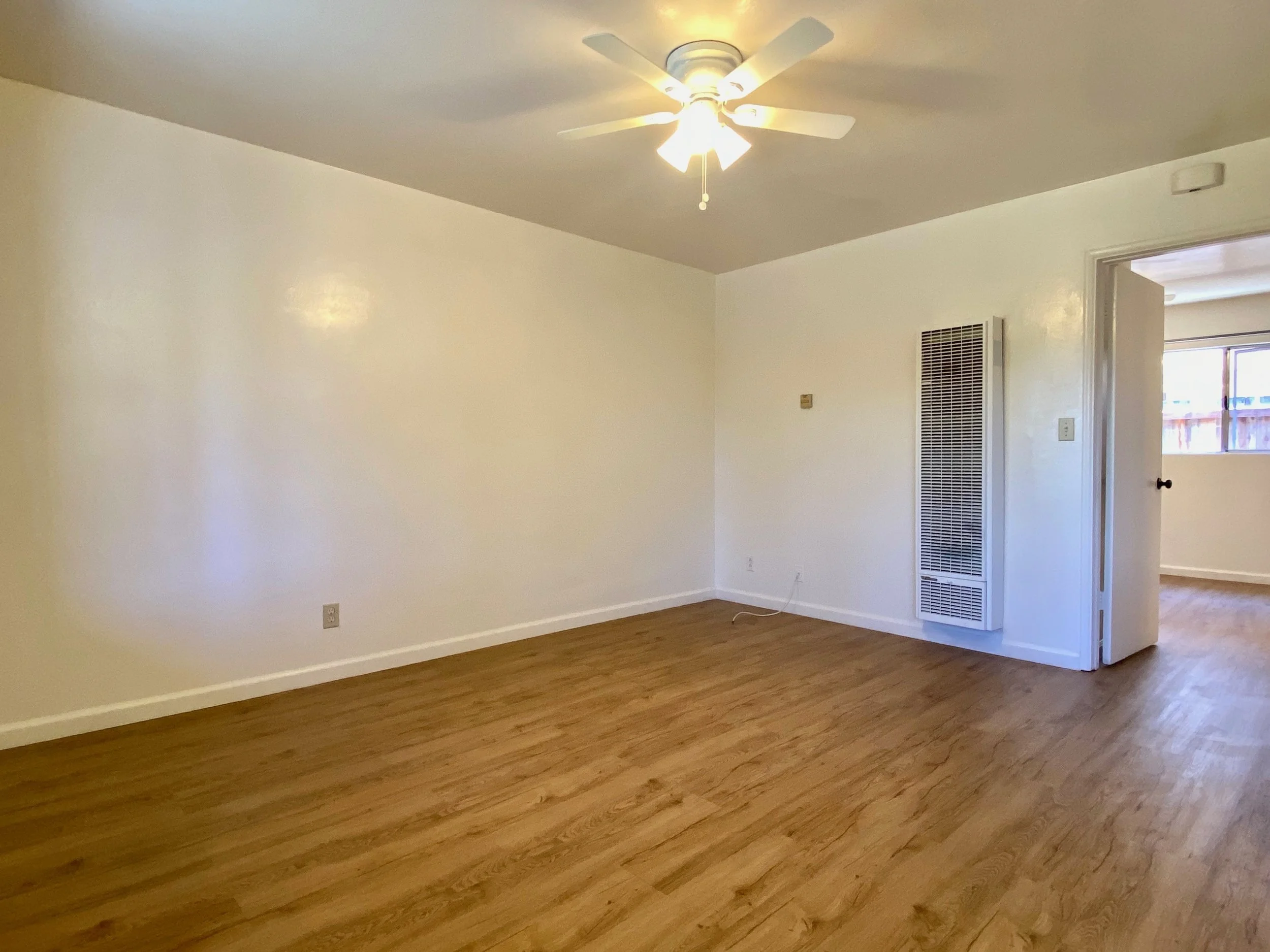 Empty living room with white walls, wooden floor, ceiling fan, and a doorway leading to another room.