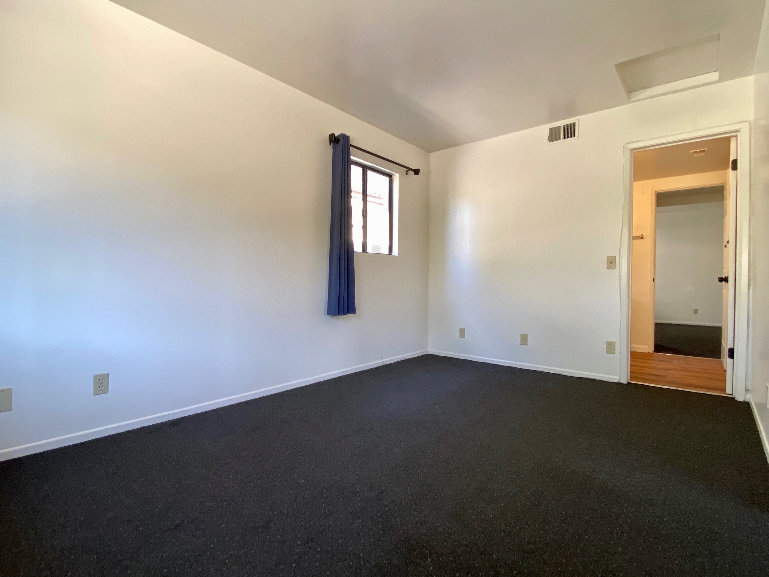 Empty room with white walls, a window with a blue curtain, a door leading to another room, and black carpeted flooring.