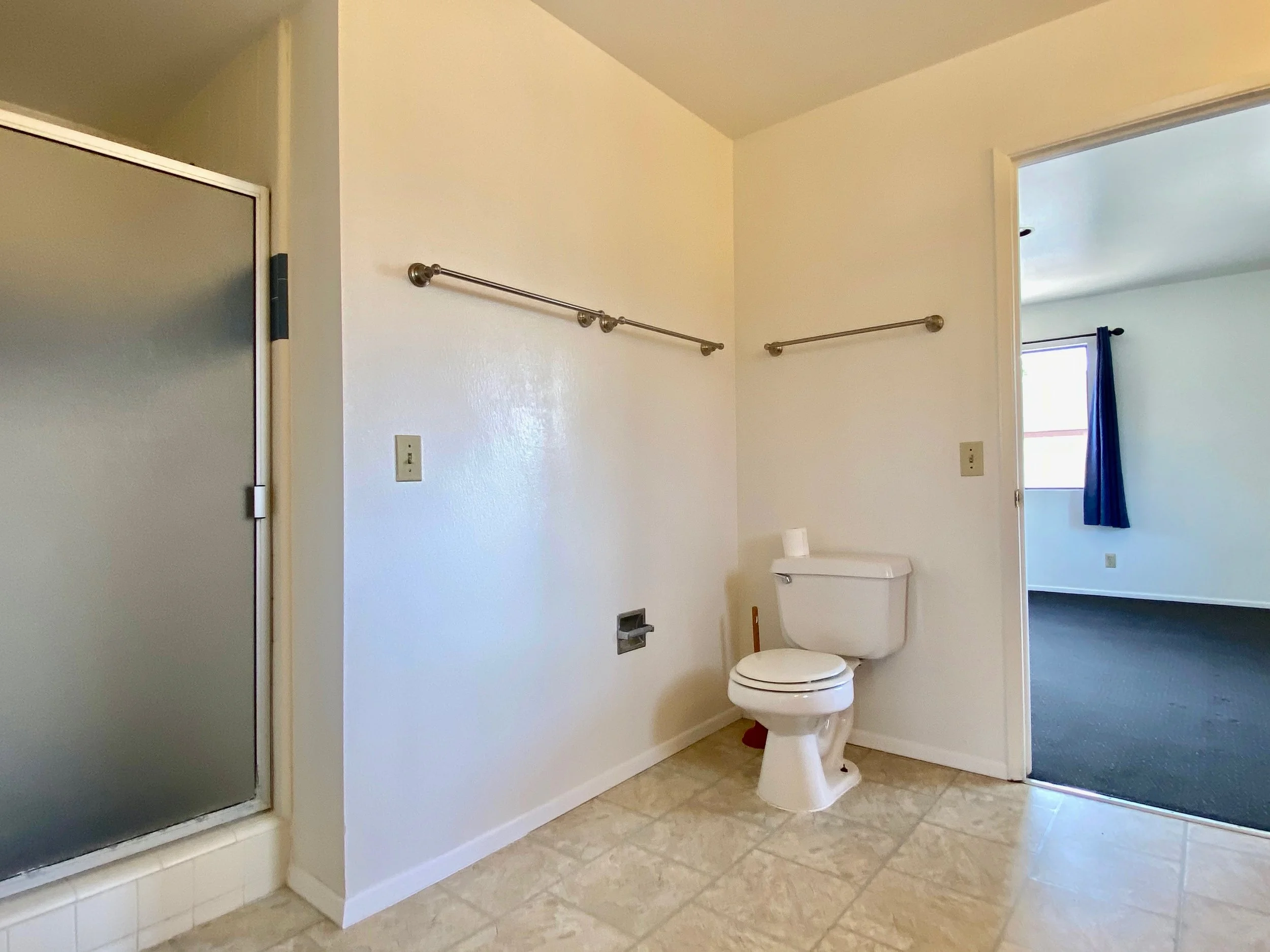 Bathroom with a toilet, two metal towel bars on the wall, a shower with a frosted glass door, a window with a blue curtain, beige tiled floor, and light-colored walls.