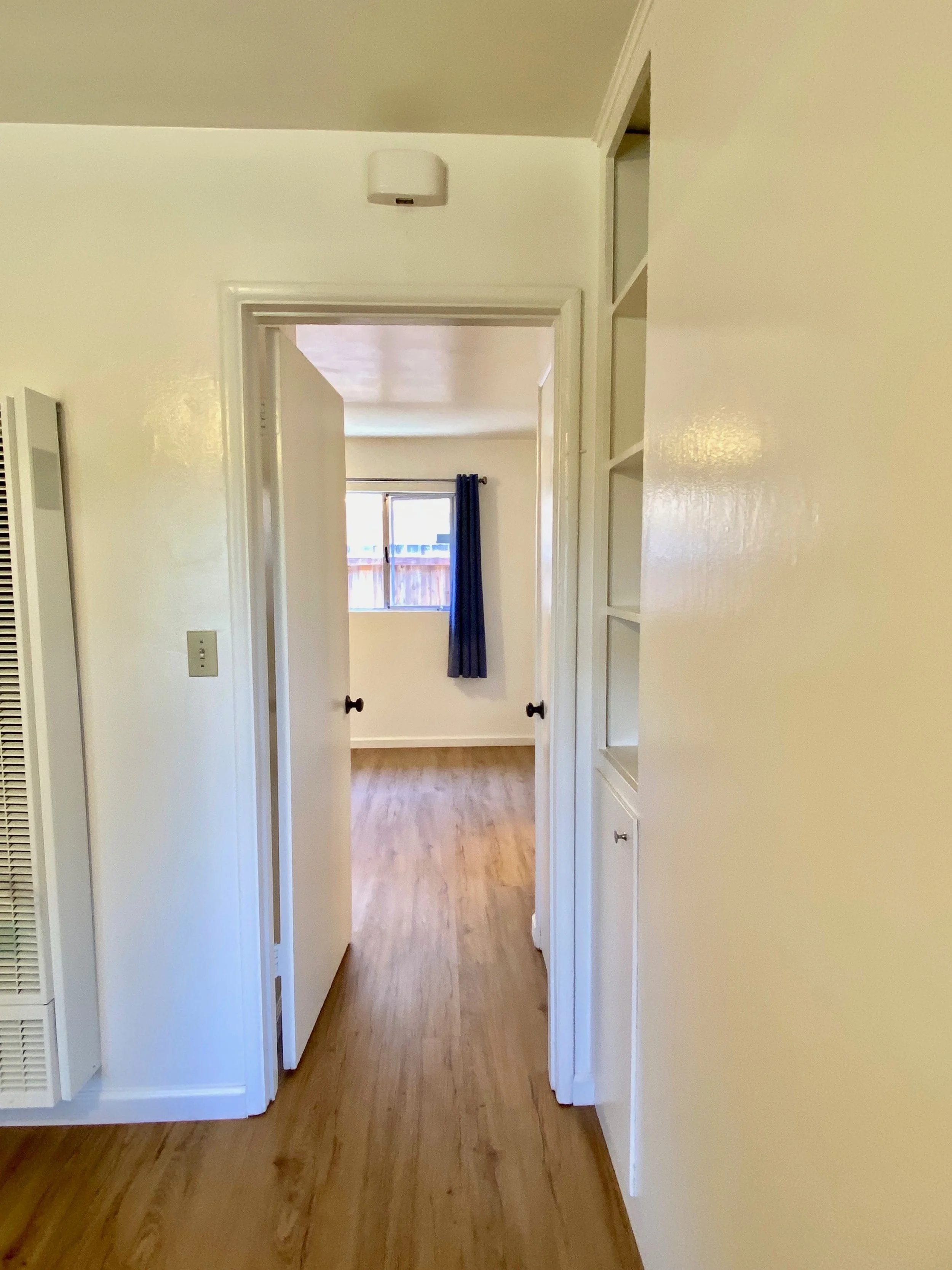 Interior view of a hallway with hardwood floors, leading to a room with a window and blue curtains, white walls, and doorways.
