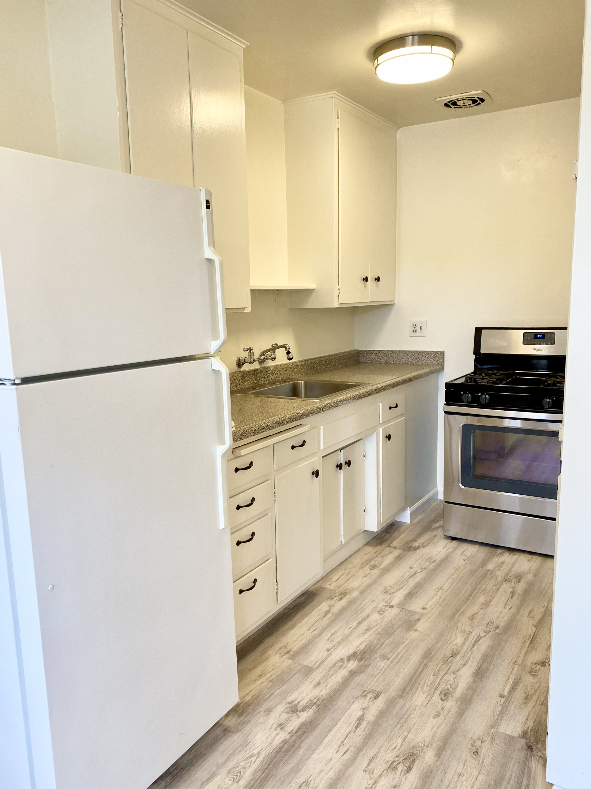 A small kitchen with white cabinets, a white refrigerator, a stainless steel stove, a gray countertop, a stainless steel sink with a gooseneck faucet, and wooden flooring.