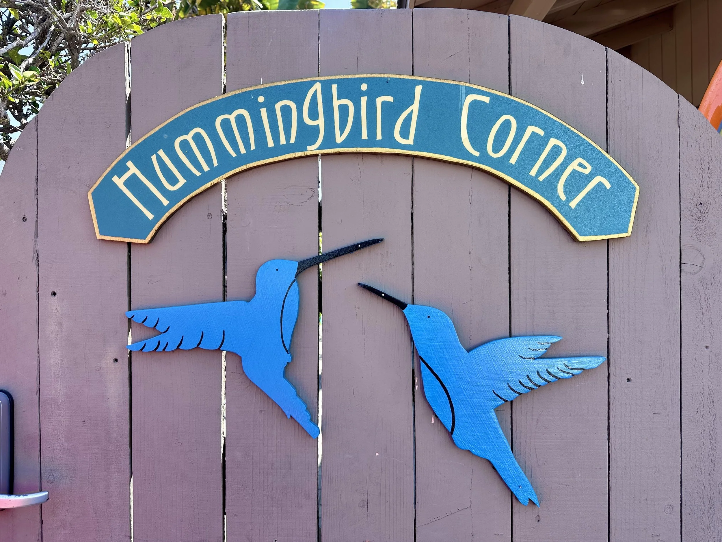 Blue decorative hanging hummingbird figures on a gray wooden fence with greenery in the background. A sign on the gate reads "Hummingbird Corner".