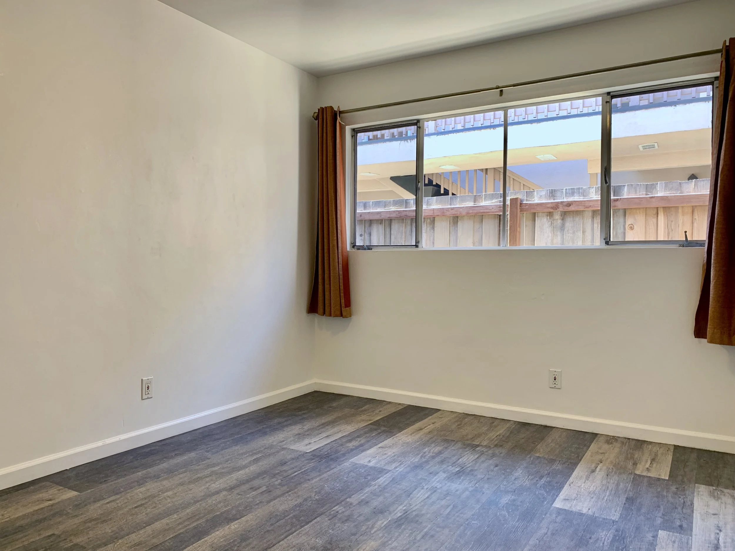 Empty room with grey wood floor, plain off-white walls, large window with curtain, and electrical outlets.