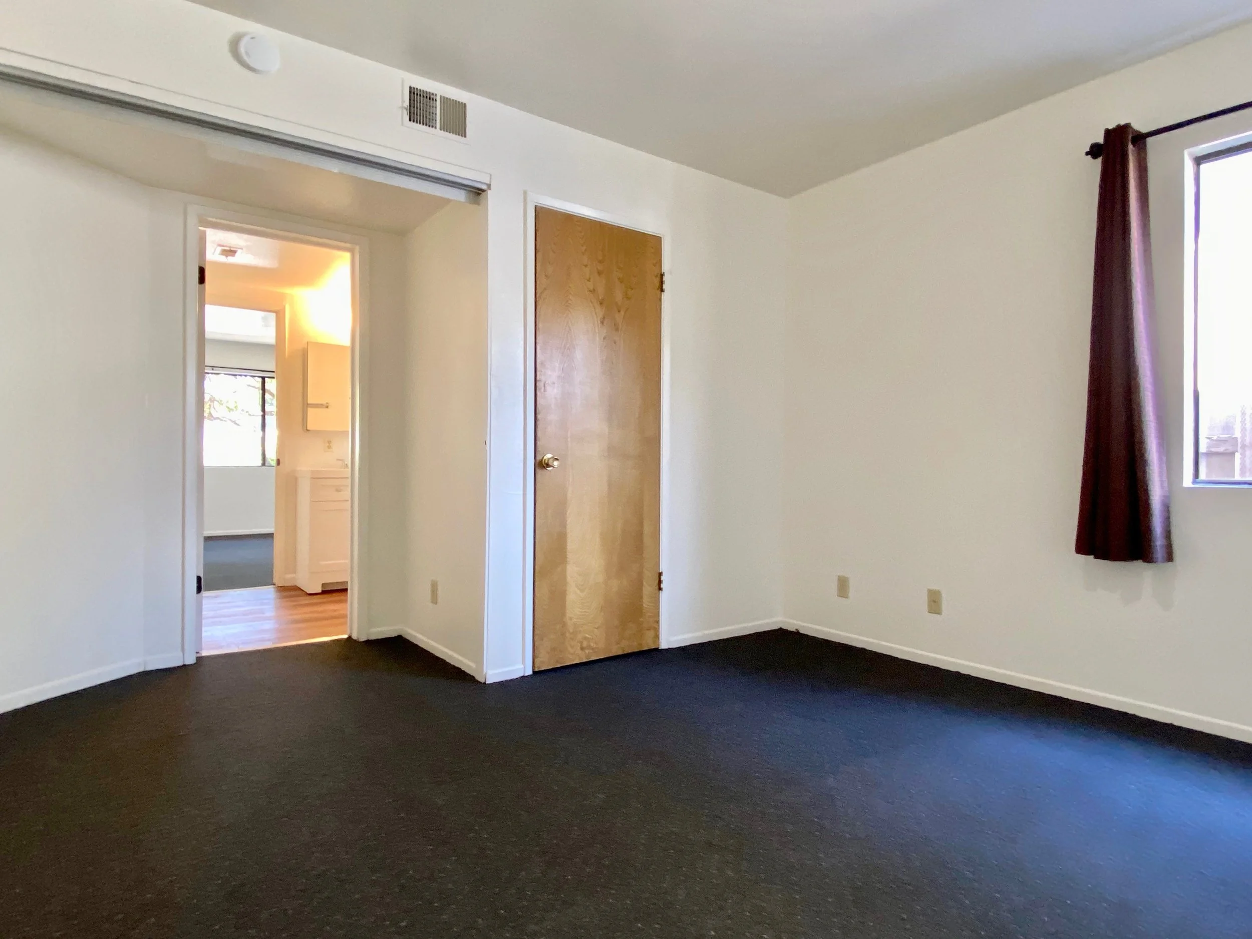 Empty room with beige walls, dark carpet, and a window with reddish-brown curtains. There is an open doorway leading to another room with hardwood flooring and a window.
