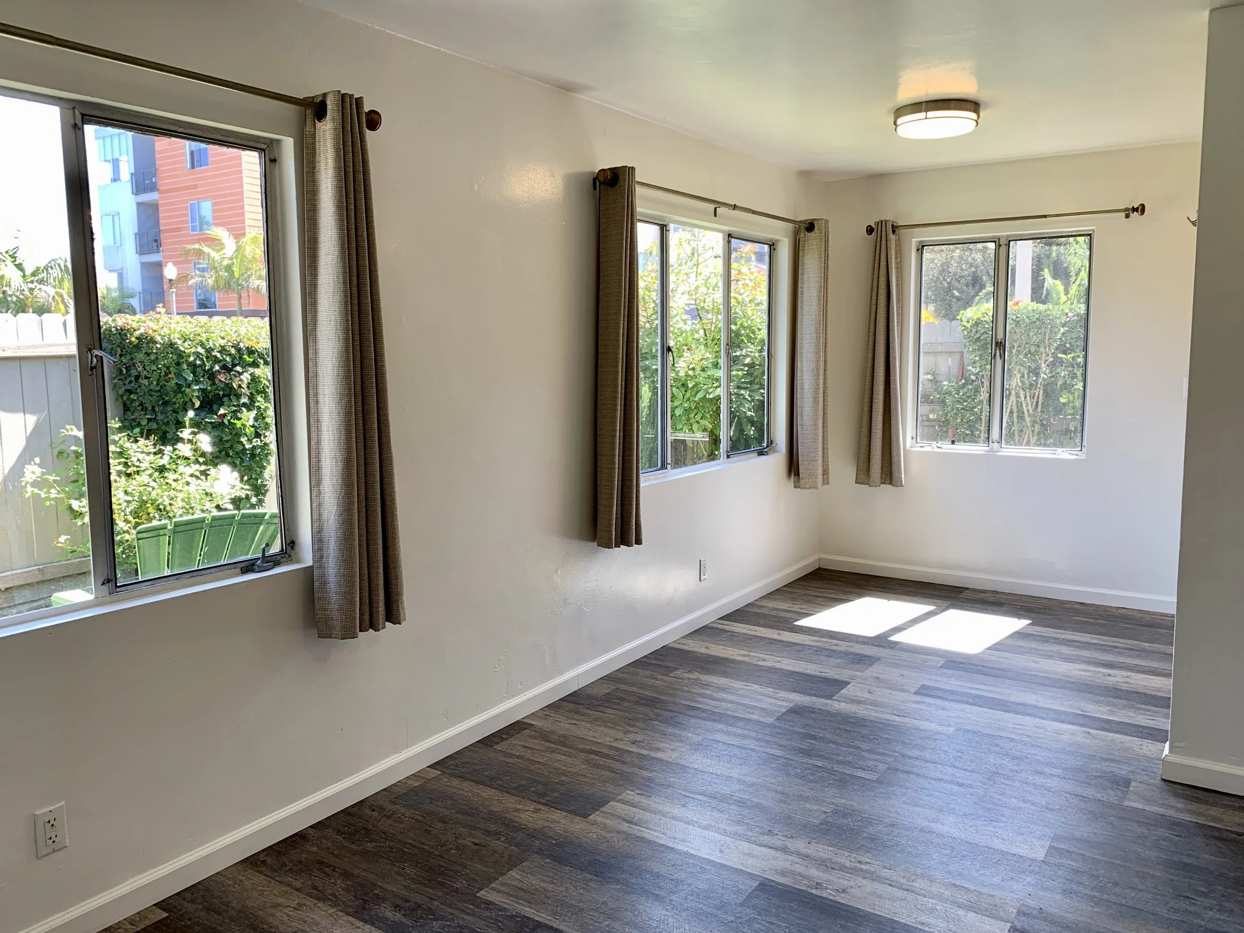 Empty room with three large windows, beige curtains, a white ceiling, a modern ceiling light fixture, brown wood-look flooring, and a white wall with an electrical outlet.