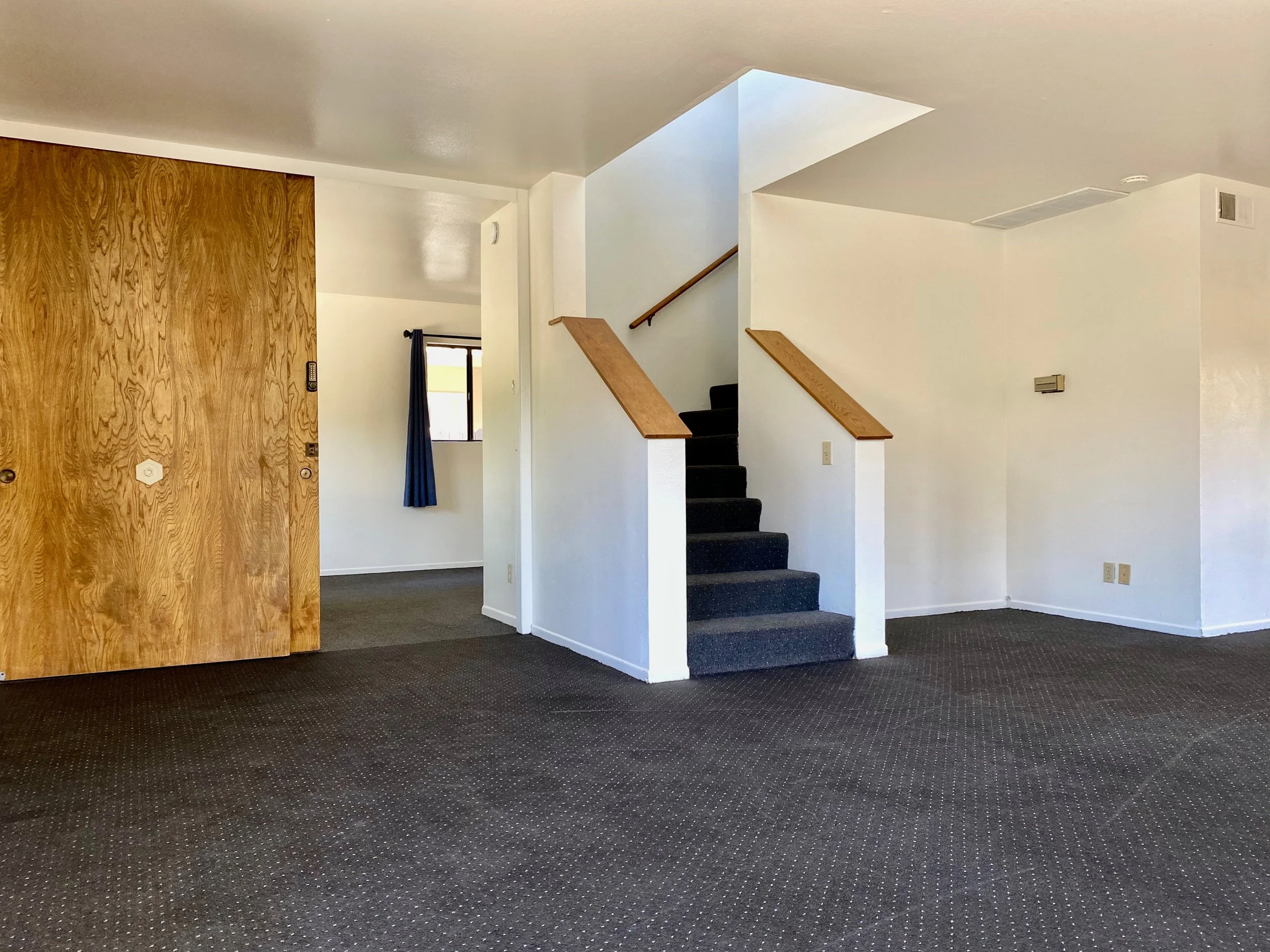 Interior of a residential or office space with carpeted floor, white walls, a staircase with black carpet and wooden handrails, a wooden door, window with blue curtains, and a ceiling vent.