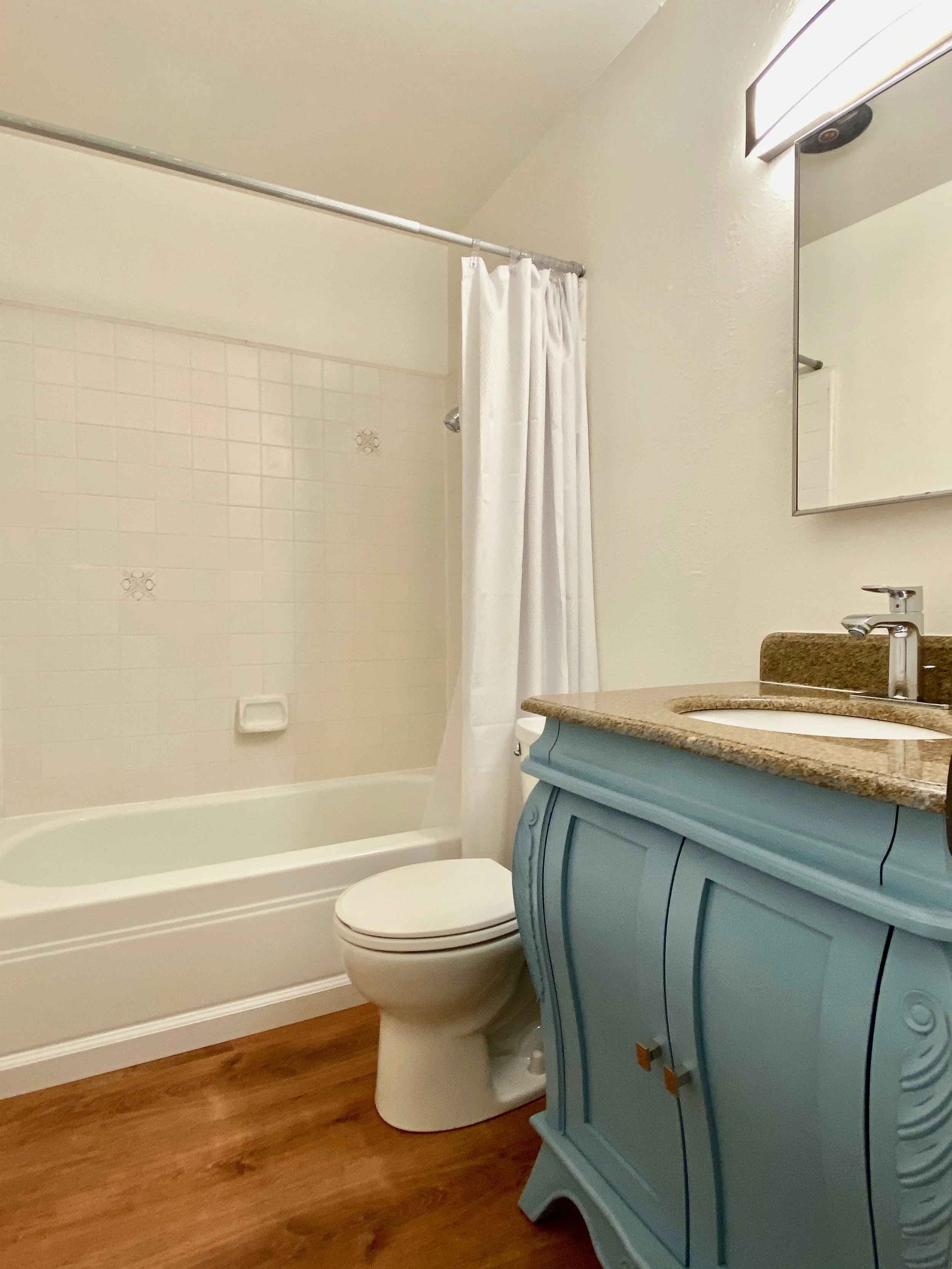 A bathroom with a bathtub and a shower with a white curtain. There is a beige countertop with a sink, a mirror above it, and a blue vanity with decorative carvings. The bathtub has a soap holder and the toilet is next to the vanity. The floor is made