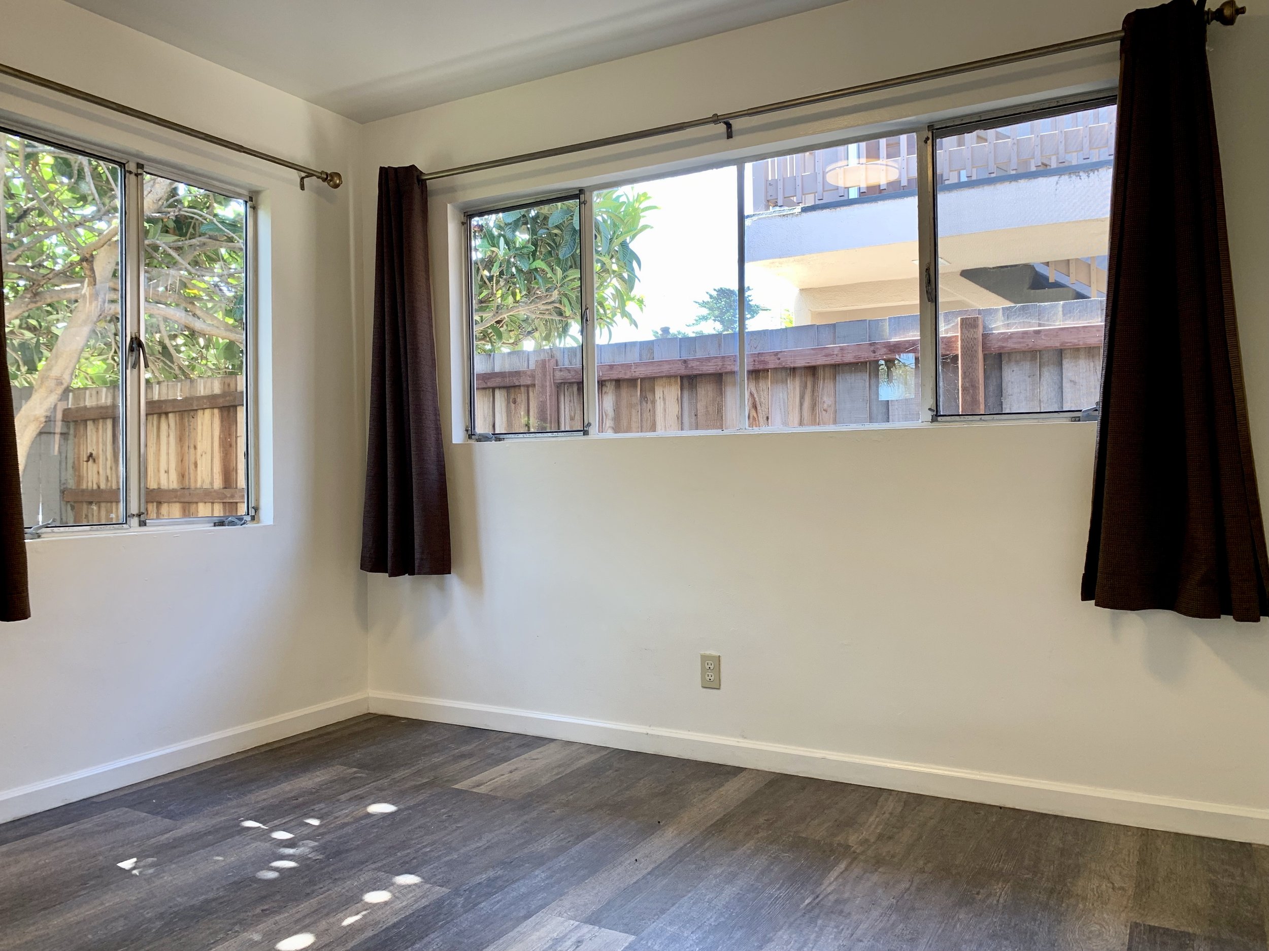 Empty room with large windows, brown curtains, white walls, dark wood flooring, and sunlight creating circular highlights on the floor.
