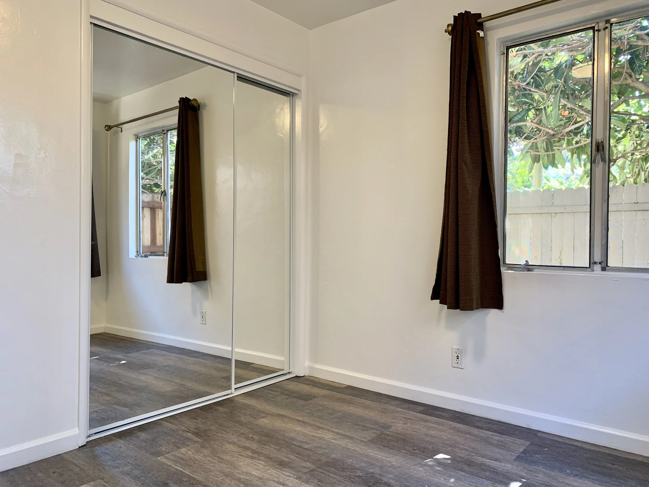 Empty room with white walls, a closet with mirrored sliding doors, and two windows with brown curtains showing green trees outside.
