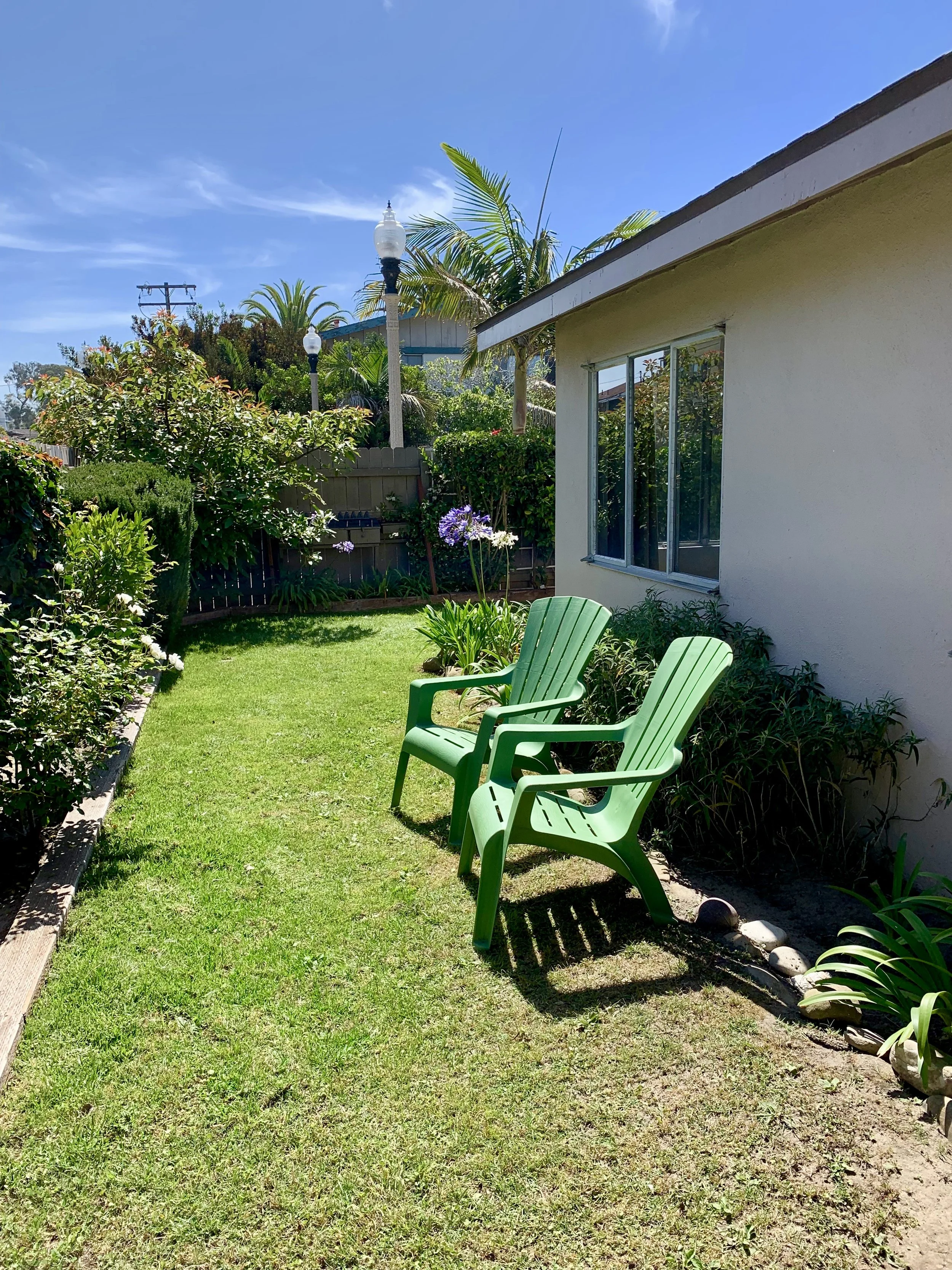 A backyard with two green plastic chairs on a grass lawn, surrounded by bushes, flowers, and trees, with a building wall and windows, under a blue sky with some clouds.