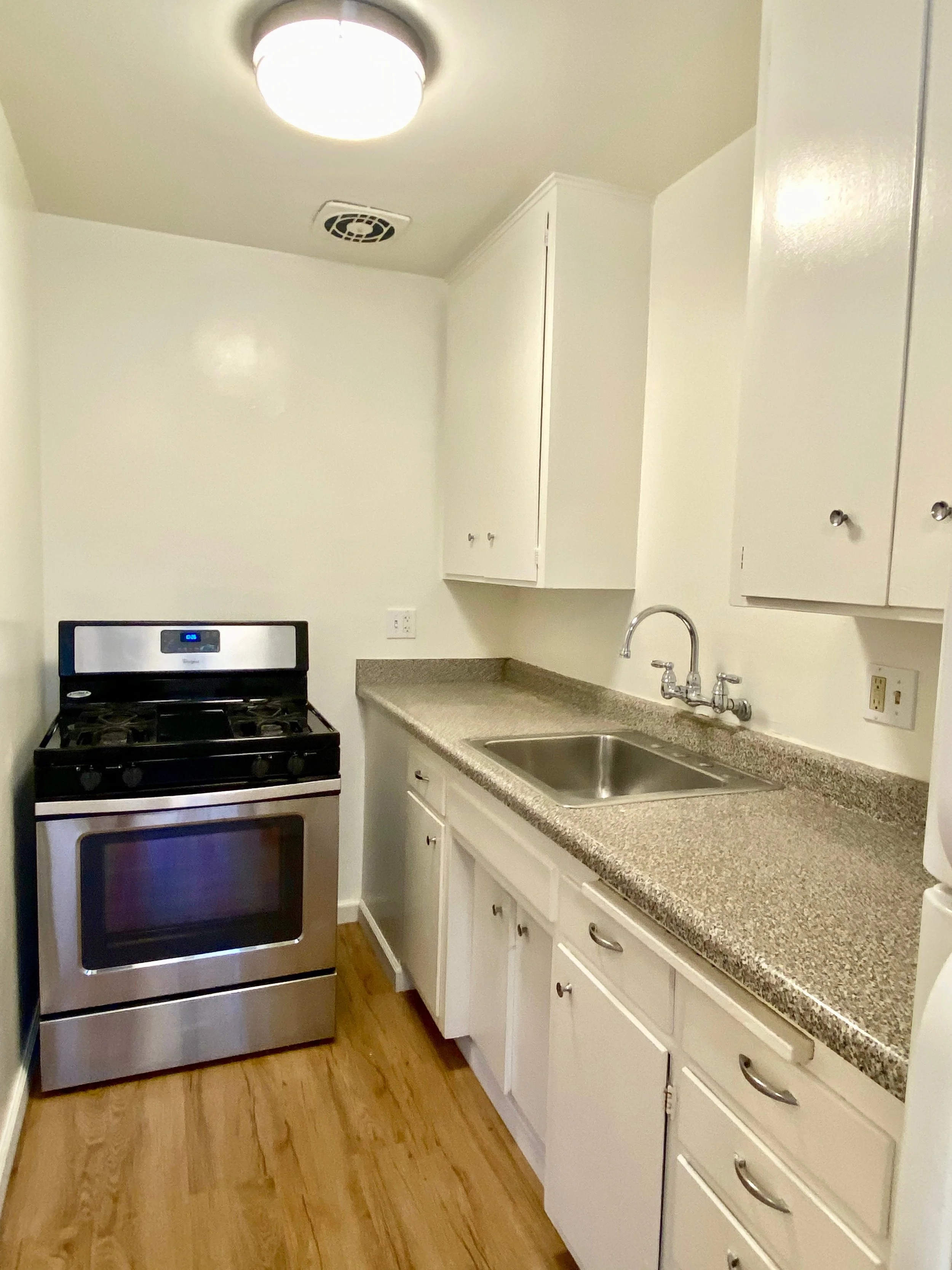 A kitchen with a stainless steel stove, white cabinets, a beige speckled countertop, a sink, and wooden flooring. There is a ceiling light and a vent on the ceiling.