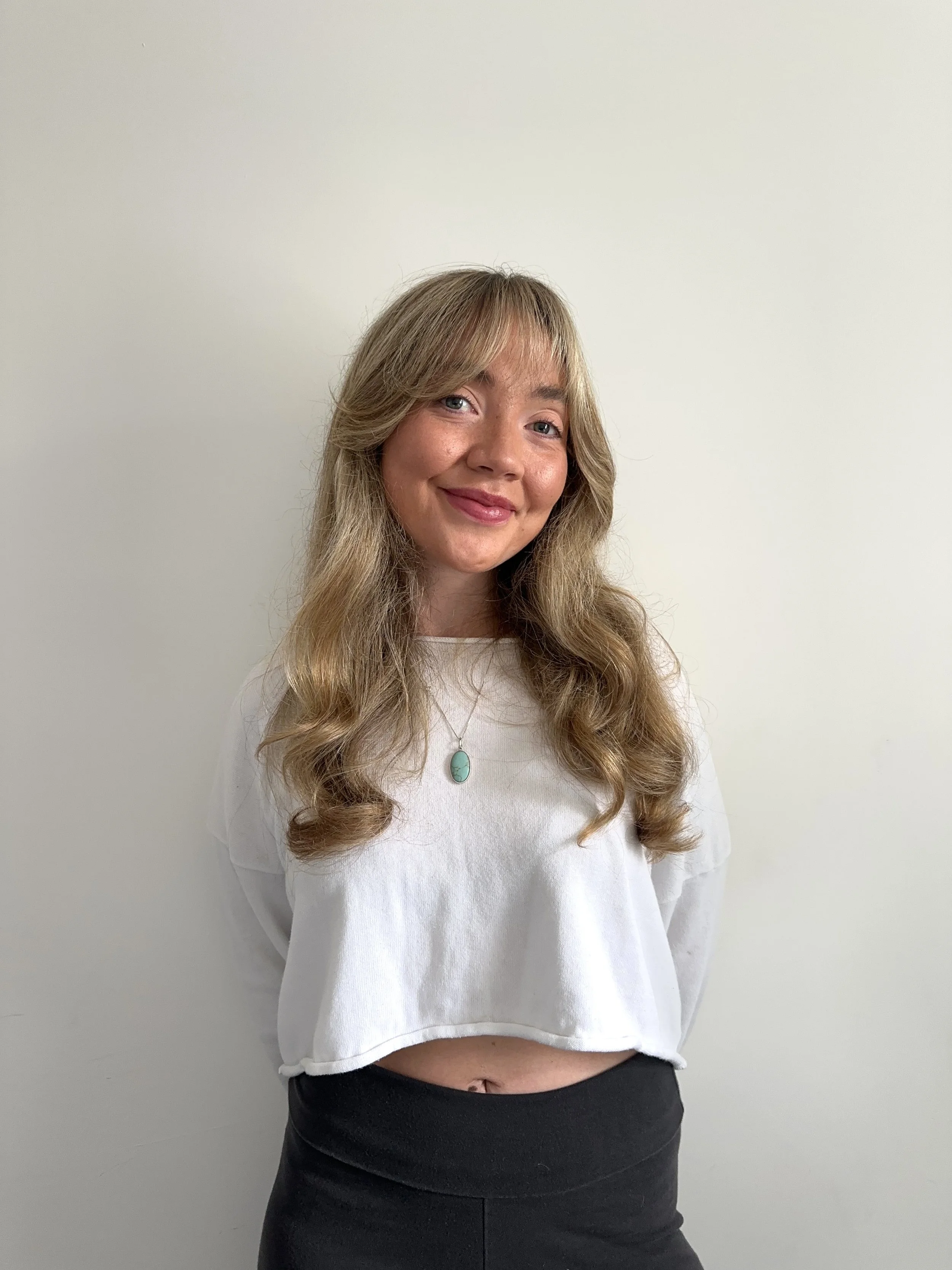 A young woman with long, wavy blonde hair and light skin smiling and looking at the camera. She is wearing a white top with a turquoise pendant necklace, and black pants. She is standing against a plain off-white wall.
