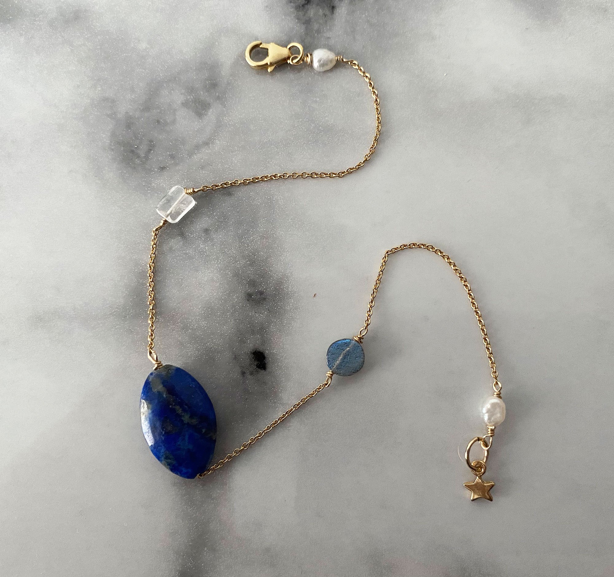 Gold chain bracelet with blue lapis lazuli stone in the centre, semi-precious stones and a star-shaped charm on a grey surface.