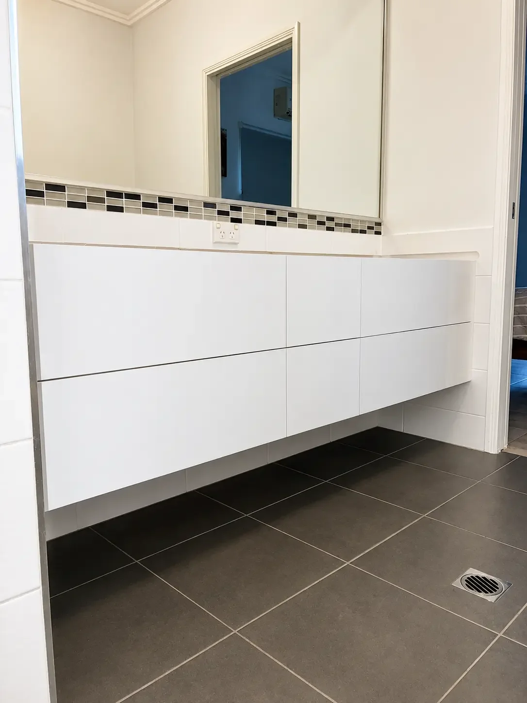 Wall-hung white bathroom vanity with handle-free drawers and dark tiled flooring.
