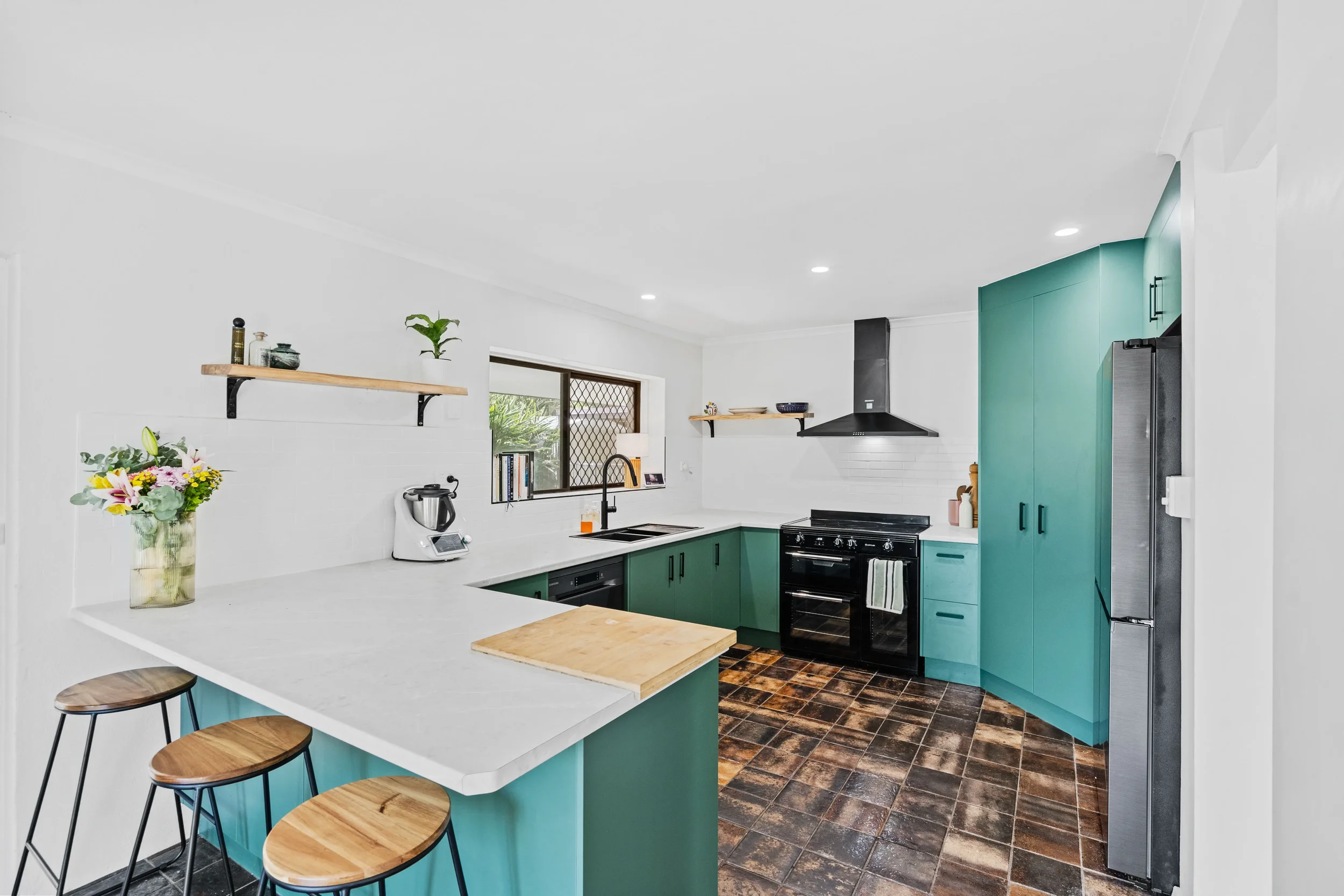 Green custom kitchen cabinetry with white benchtops, open shelving and black appliances.
