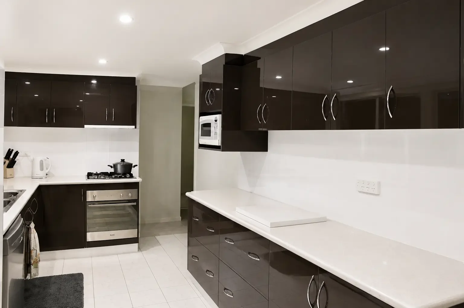 Dark kitchen cabinetry with white benchtops and built-in oven and microwave.
