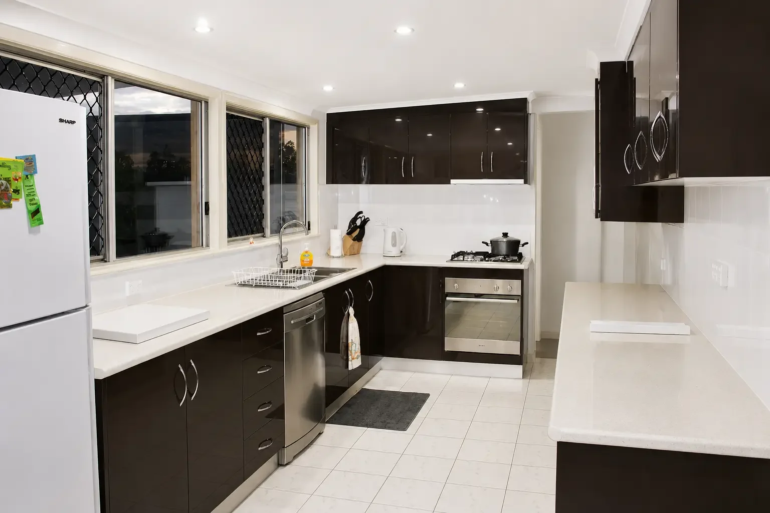 Dark kitchen cabinetry with white benchtops, gas cooktop and sink beneath the window.

