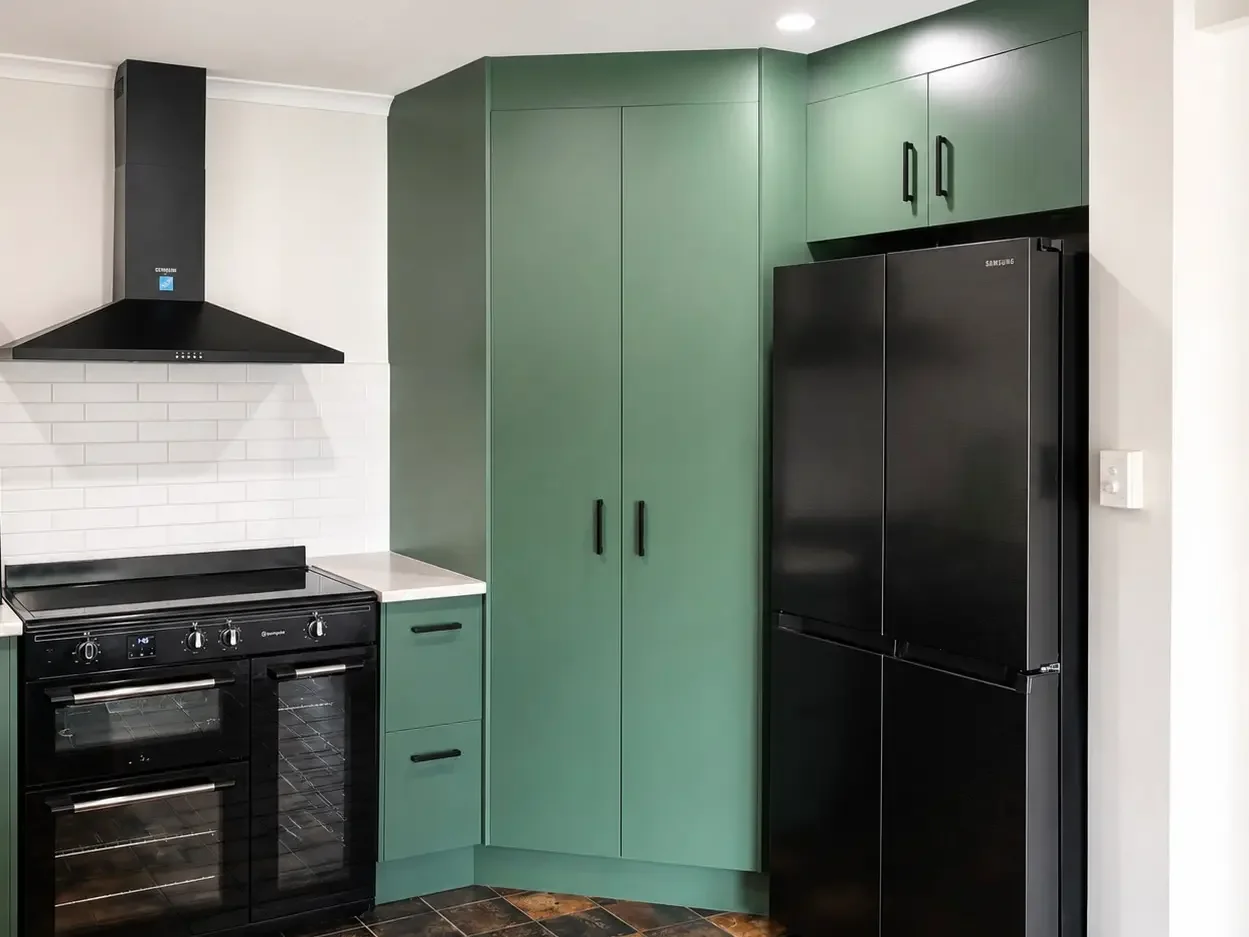 Green kitchen cabinetry with black appliances, white subway tile splashback and black rangehood.

