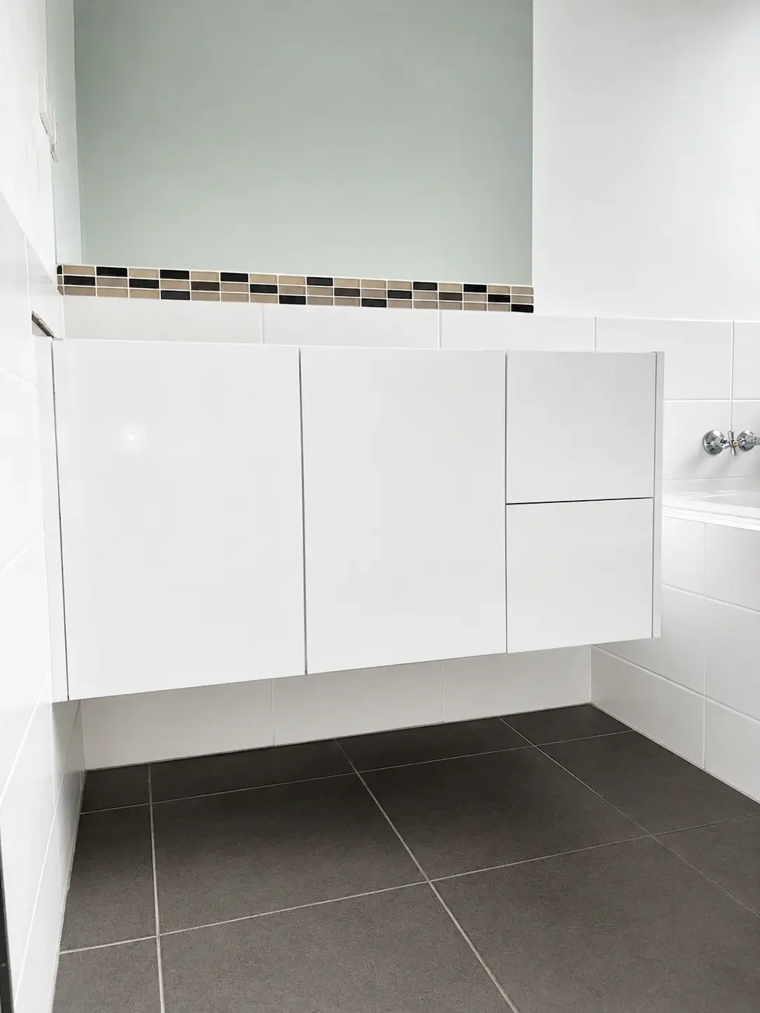 White bathroom vanity cabinetry with tiled splashback and dark tiled flooring.
