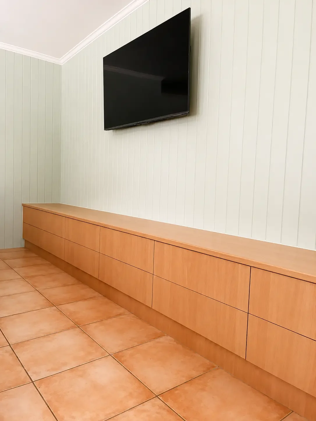 A wall-mounted flat-screen TV above a long wooden cabinet in a room with beige tiled flooring and light green vertical wood paneling on the wall.