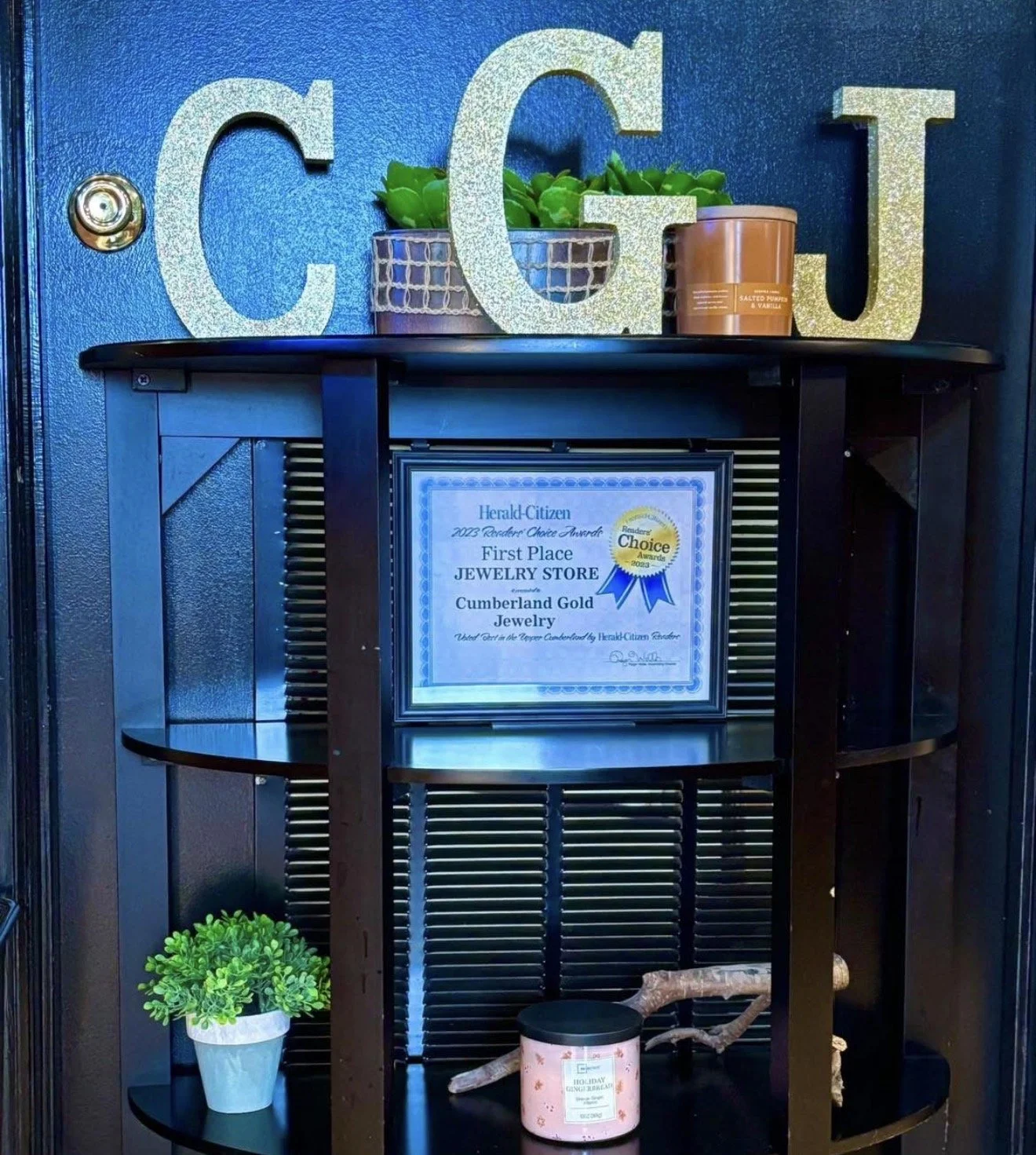 Decorative shelf with a framed certificate for first place in jewelry store, a plant, a candle, and decorative letters 'C G J' on the wall behind.
