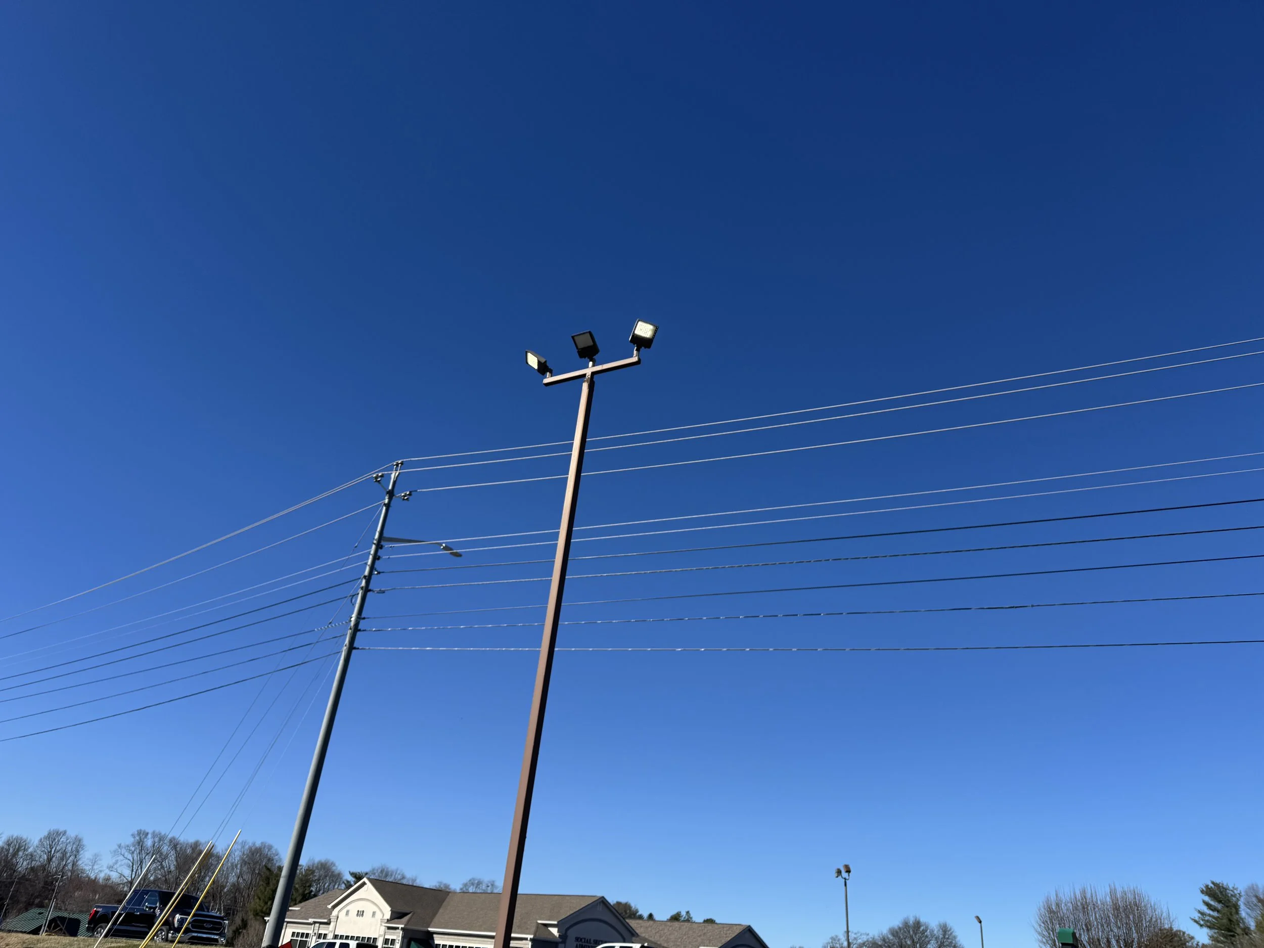A tall light pole with four floodlights against a bright blue sky. Power lines run horizontally across the image. House rooftops and trees are visible at the bottom.