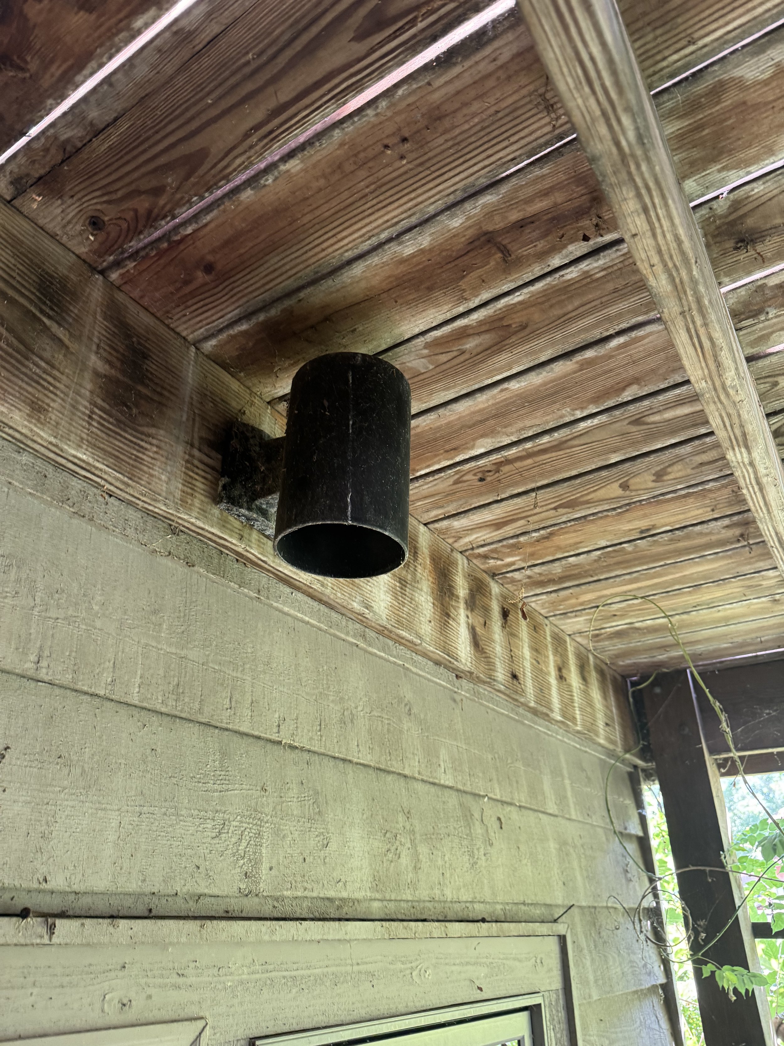 The image shows a wooden ceiling with a black pipe hanging from it, part of a porch or outdoor area with green foliage visible on the right side.