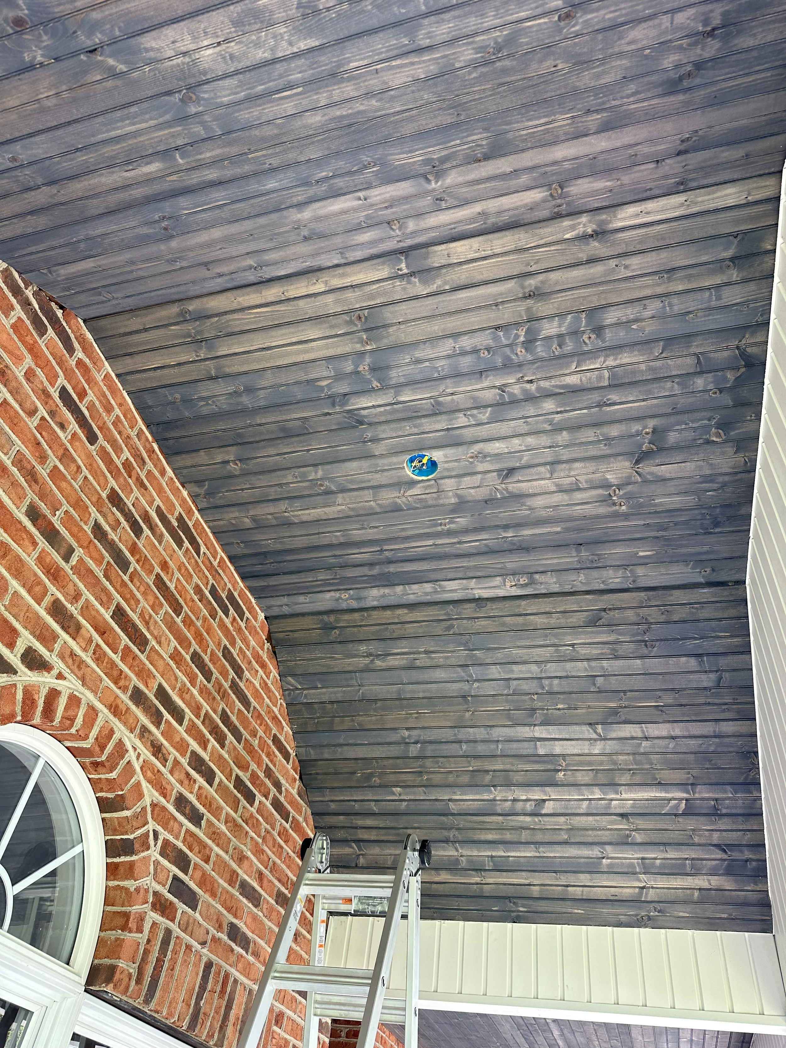 A house porch with red brick walls, a window, and a new wooden ceiling being installed with a ladder placed underneath.