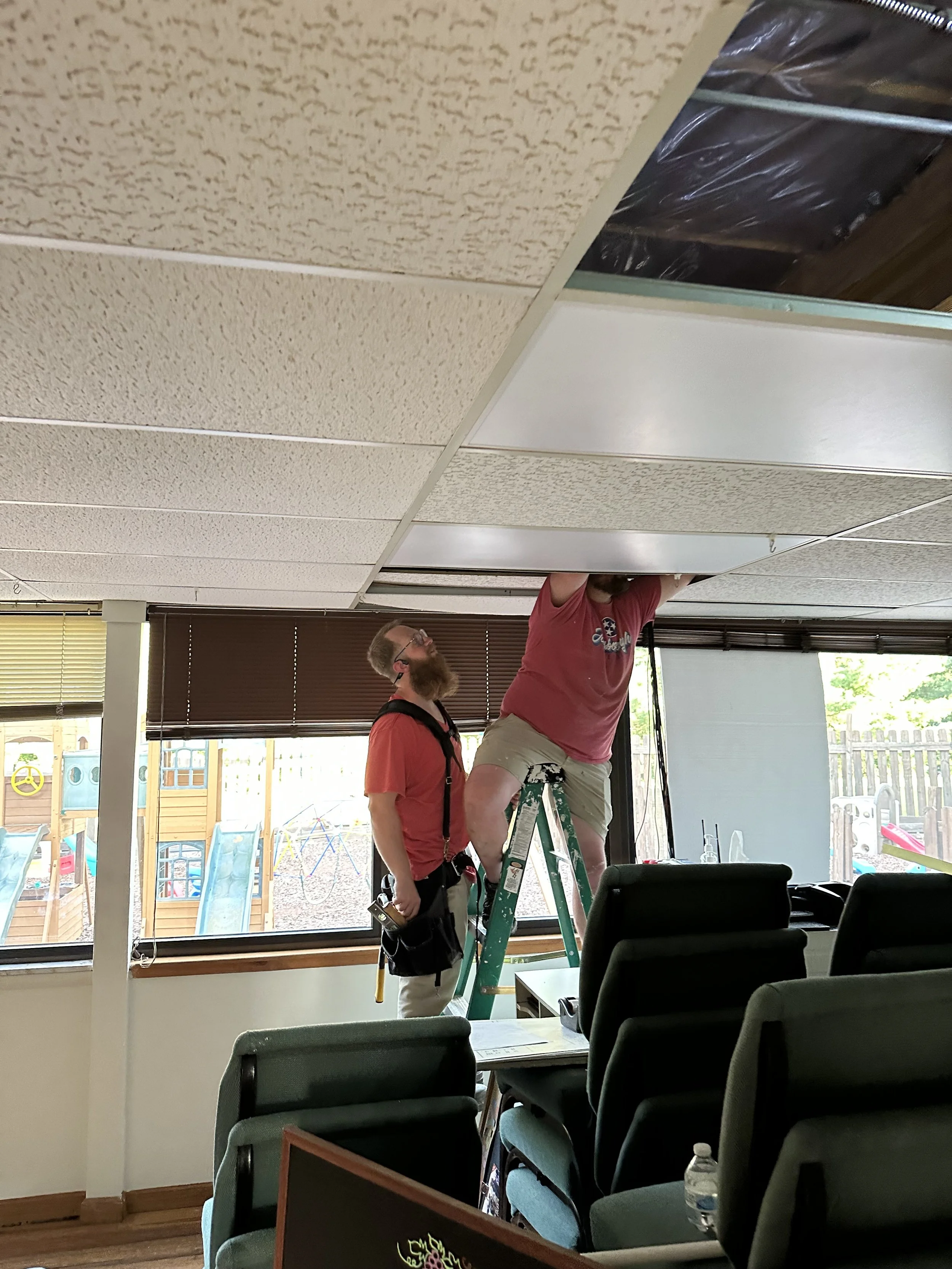 Two men working on ceiling tiles in a room with rows of chairs, window blinds, and a playground visible outside.