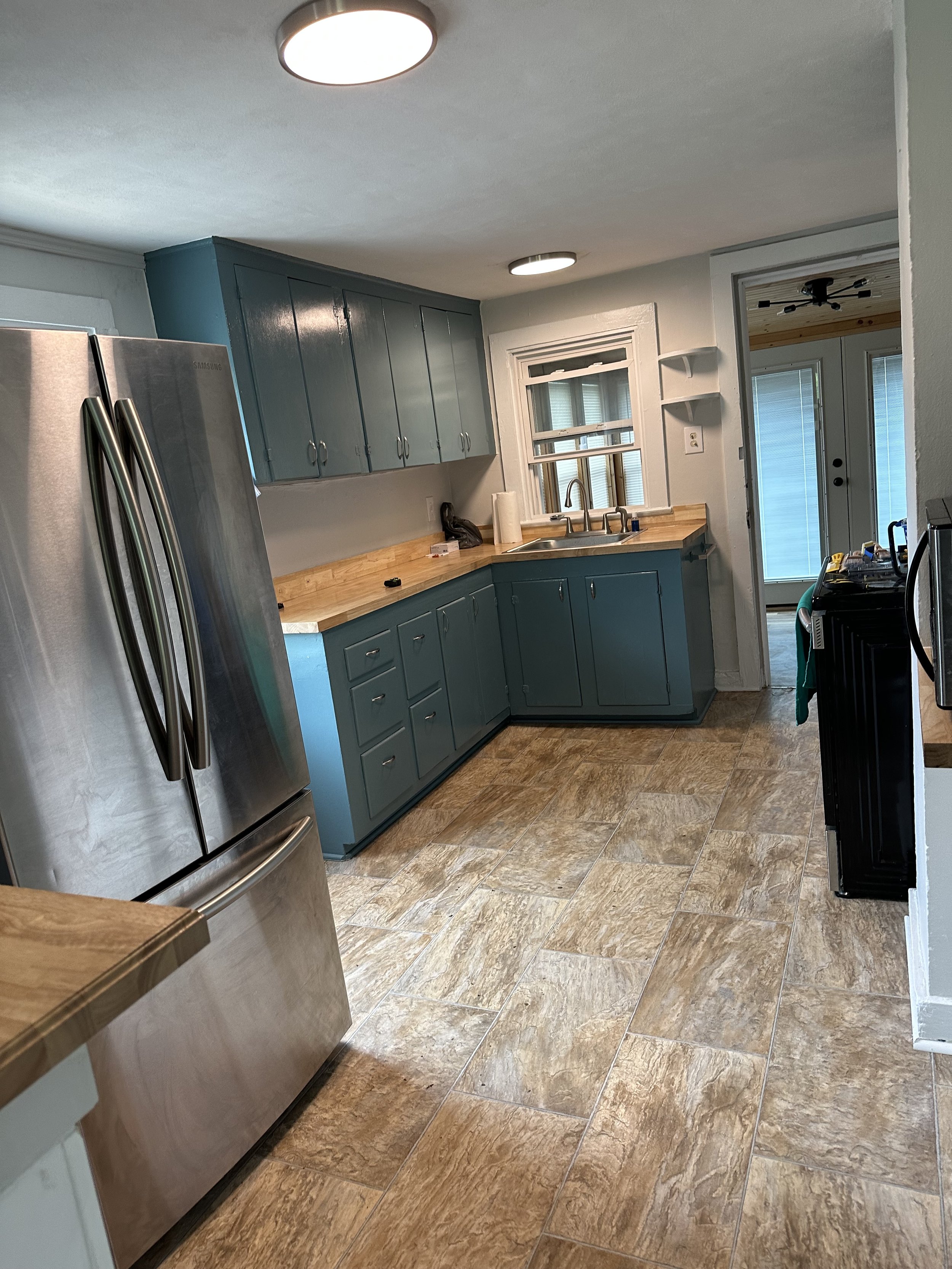 A kitchen with teal upper and lower cabinets, a wood countertop, a stainless steel side-by-side refrigerator, a window above the sink, and a door leading outside. There are ceiling lights, a paper towel holder, and some small items on the counter.