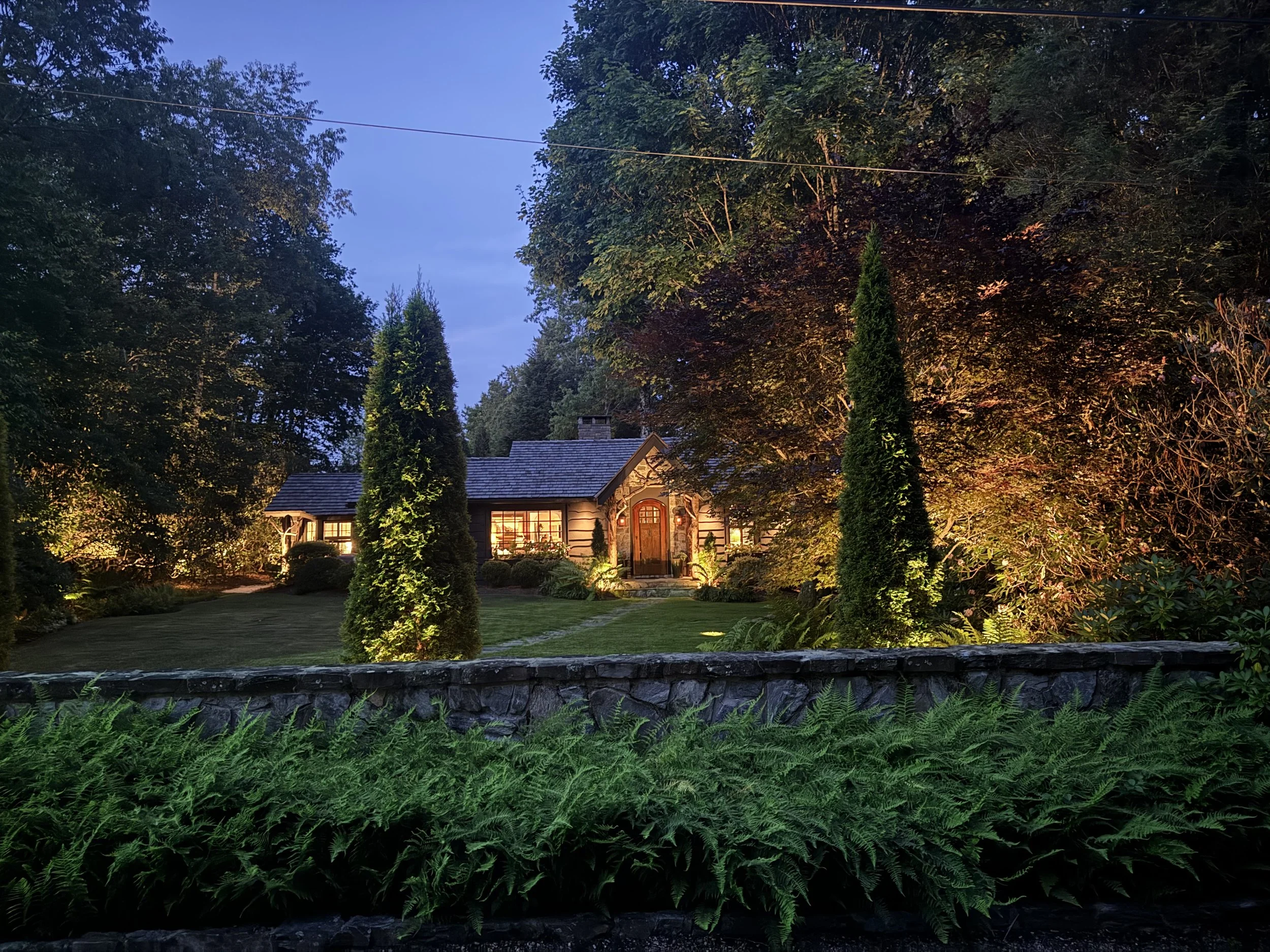 View of a house at sunset surrounded by trees and greenery, illuminated by outdoor lighting.