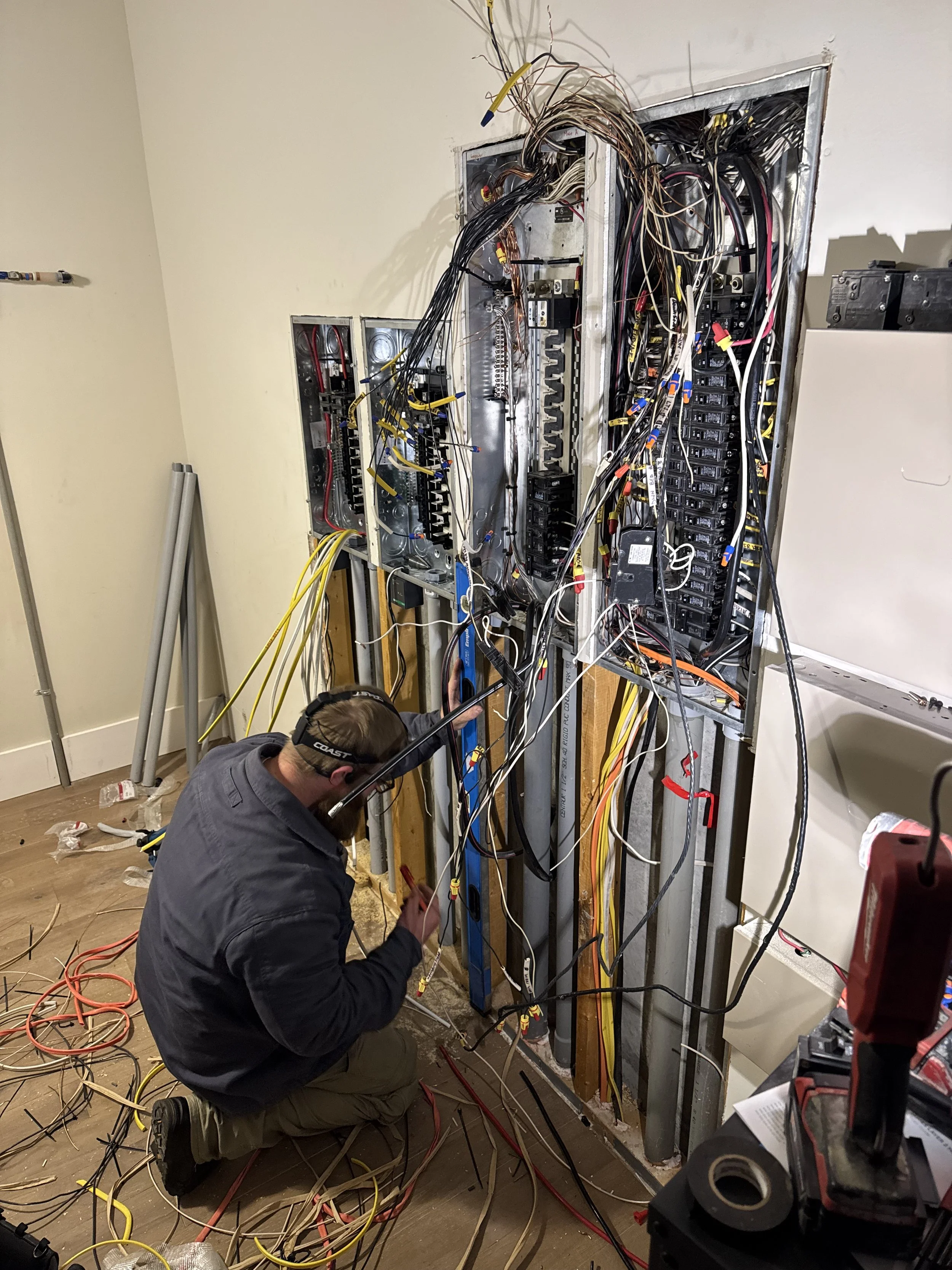 A person wearing glasses and a headset works on electrical wires behind an open wall panel in a room. The floor is scattered with wires and tools.