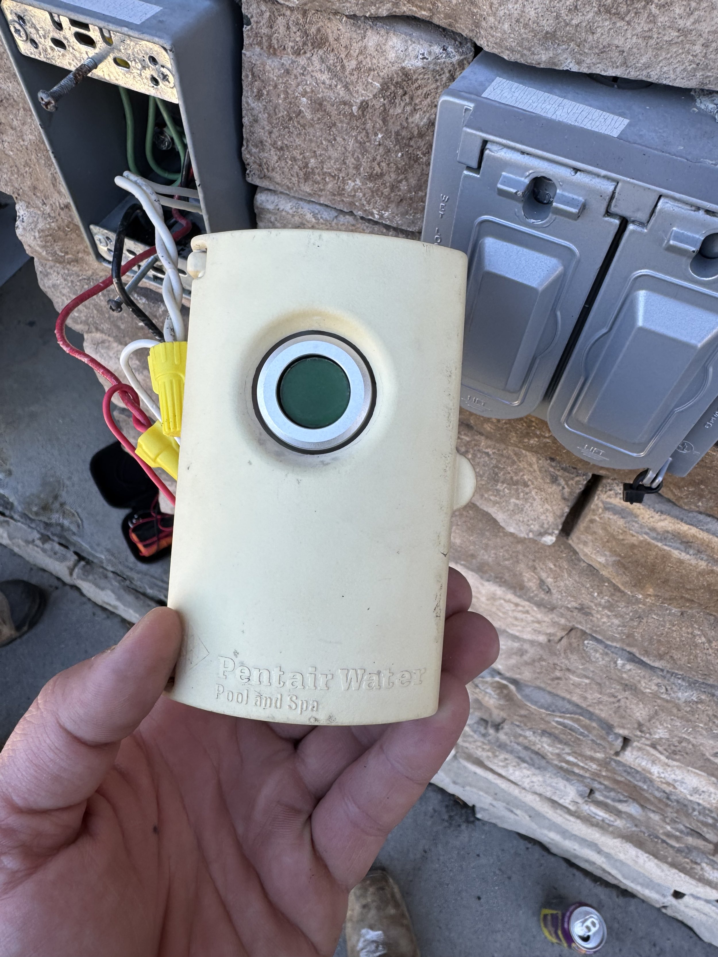 Person holding a beige remote control labeled 'Pentair Pool and Spa' with electrical wiring and metallic boxes behind, mounted on a stone surface.