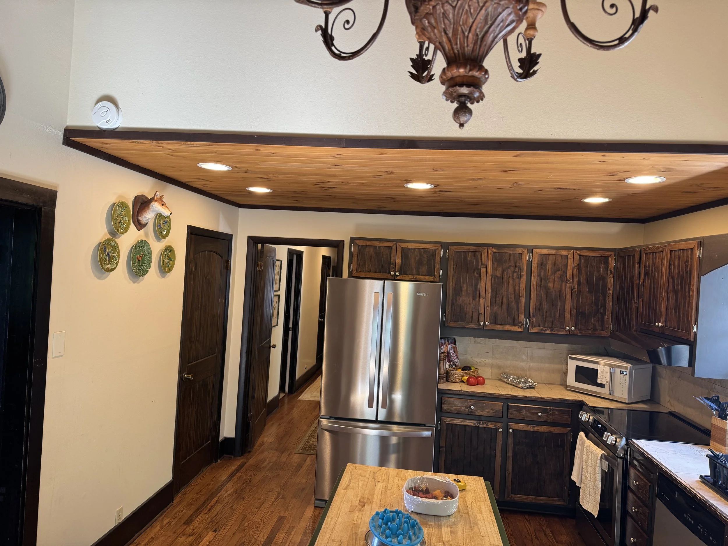 A kitchen with dark wood cabinets, a stainless steel refrigerator, and a countertop with apples and a microwave. The ceiling has wooden paneling and recessed lighting. Decor includes a wall-mounted deer head and yellow ceramic plates with nature scen