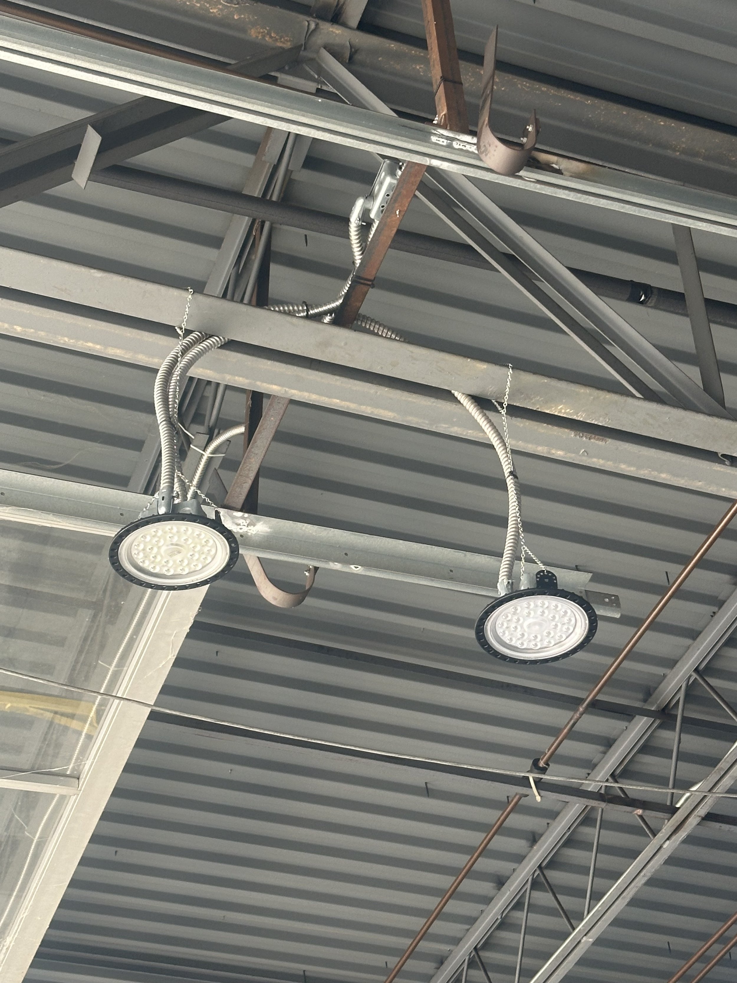 Ceiling of an industrial building with metal beams, electrical conduits, and two LED ceiling lights.