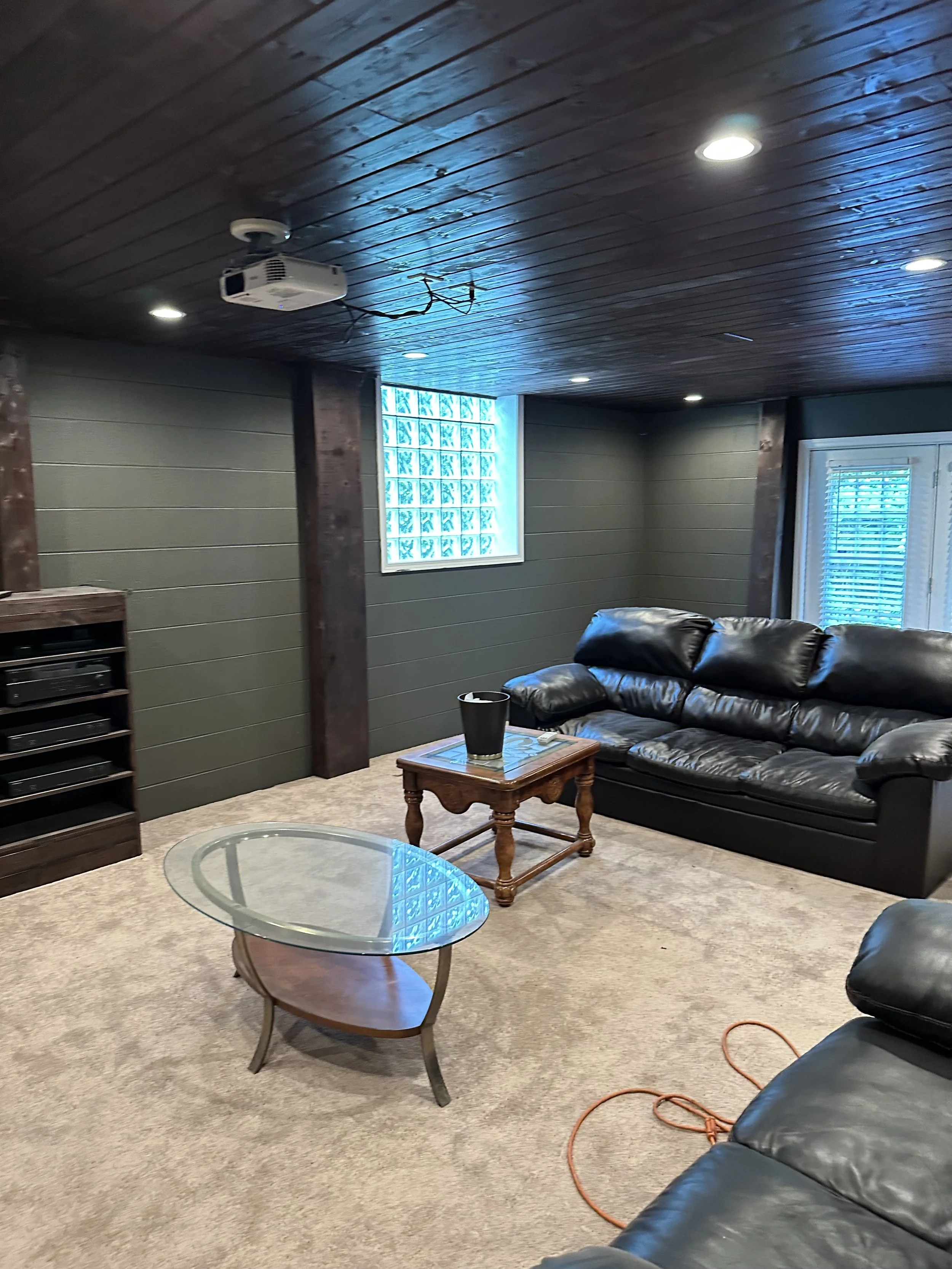 Living room with a black leather sofa, a glass coffee table, a wooden side table, a window with blinds, a wall-mounted projector, a satellite receiver, and ceiling lights.