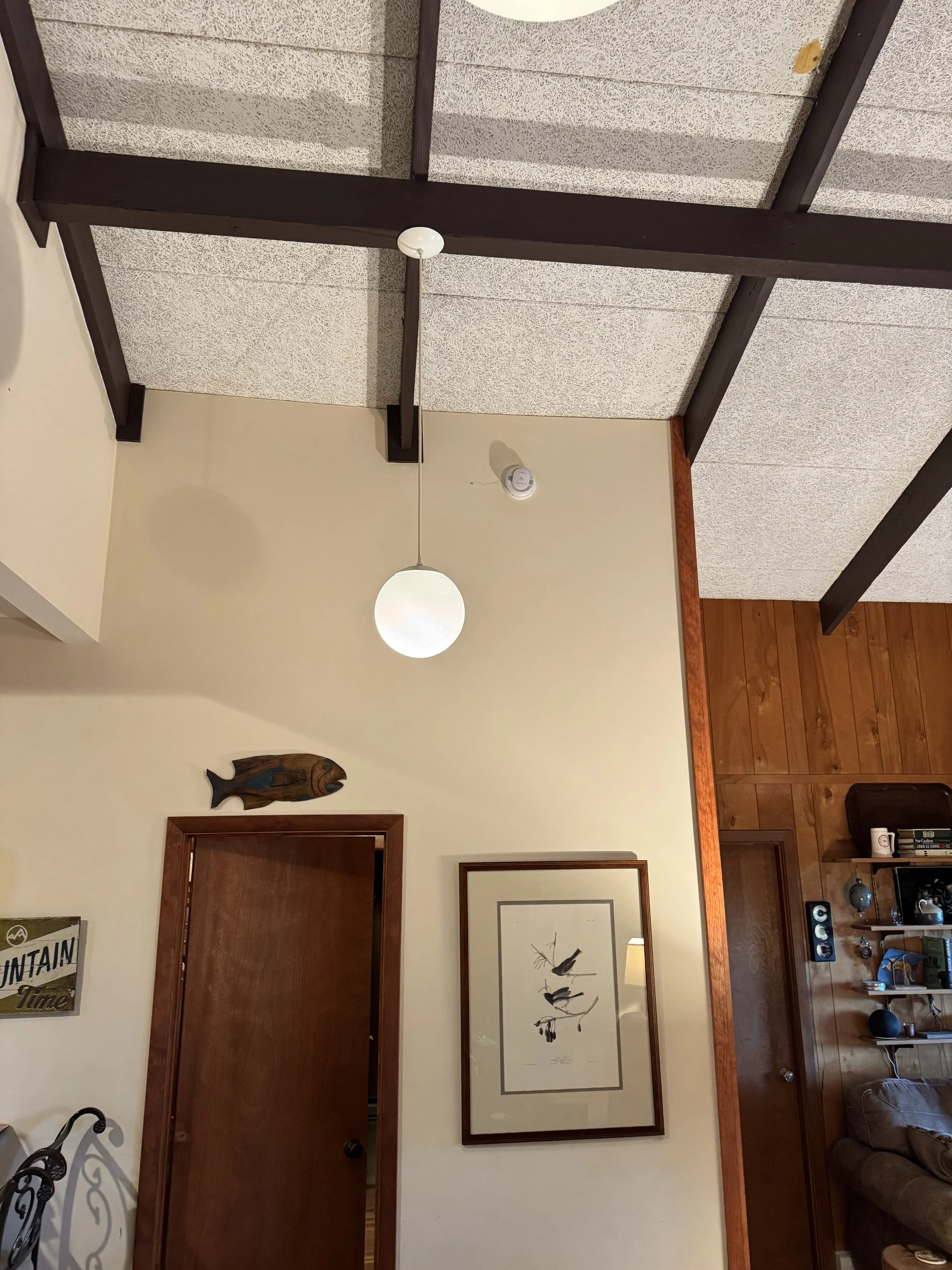 Living room with beige walls, framed bird artwork, wooden door, and dark wood beam ceiling. There is a living room with wooden flooring, a second framed picture, and multiple small decorative objects on a shelf.