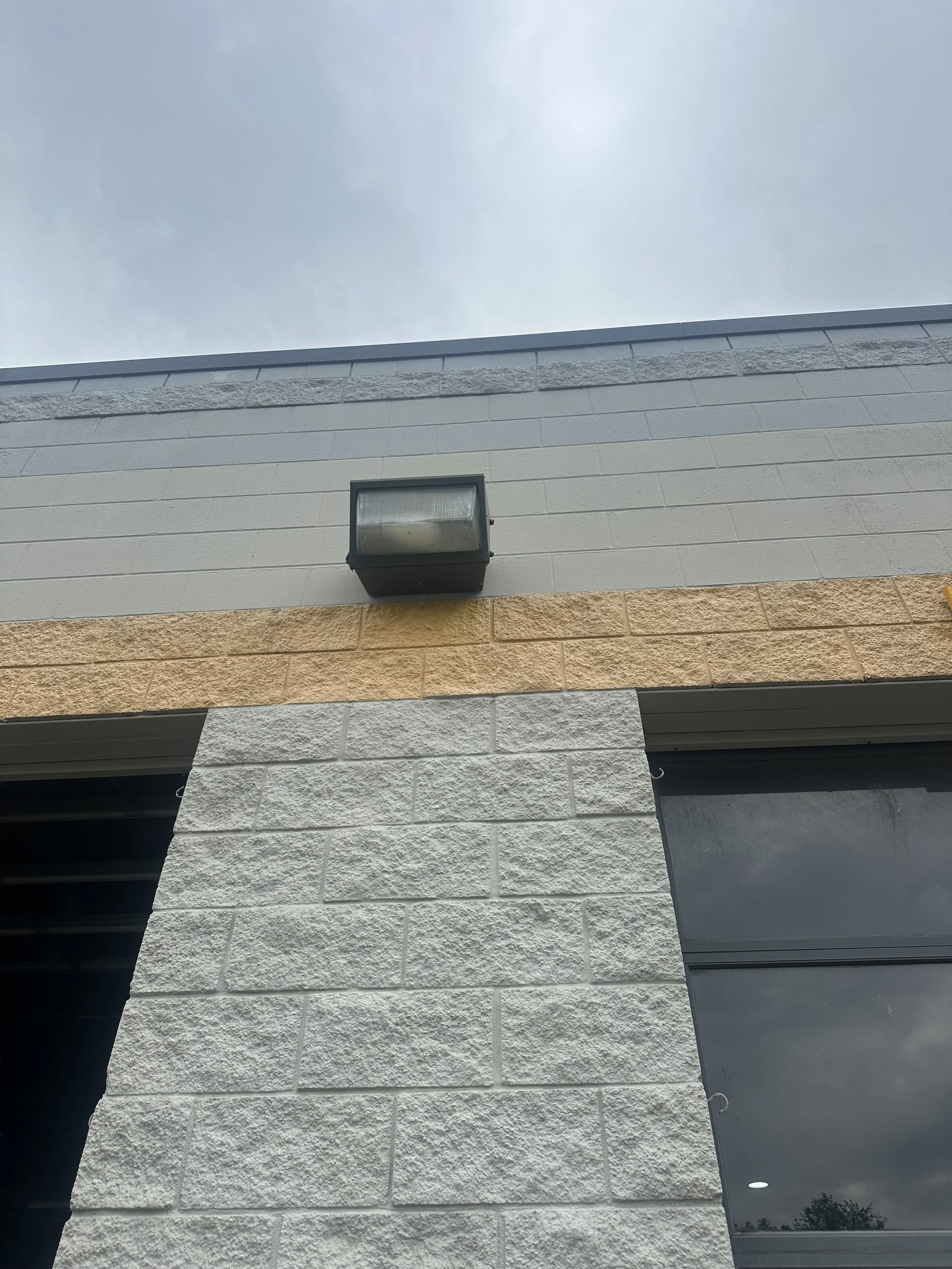 Part of a building exterior with different textured and colored wall panels, a black outdoor light fixture, and a reflected sky with some clouds.
