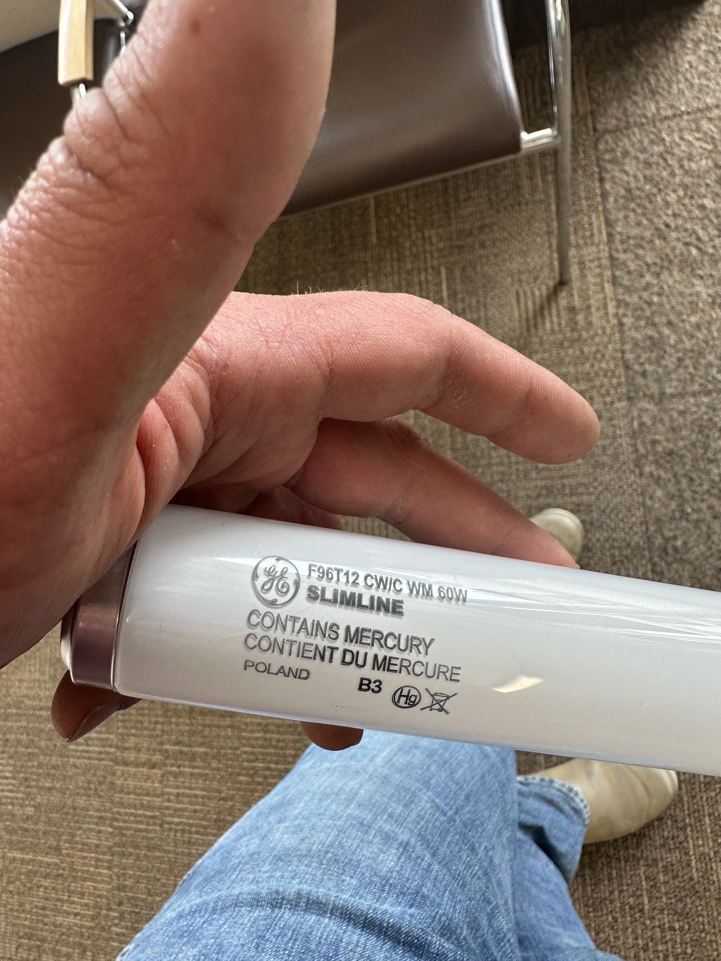 Close-up of a person holding a mercury-containing light bulb with safety warnings, against a background of a beige chair and carpeted floor.