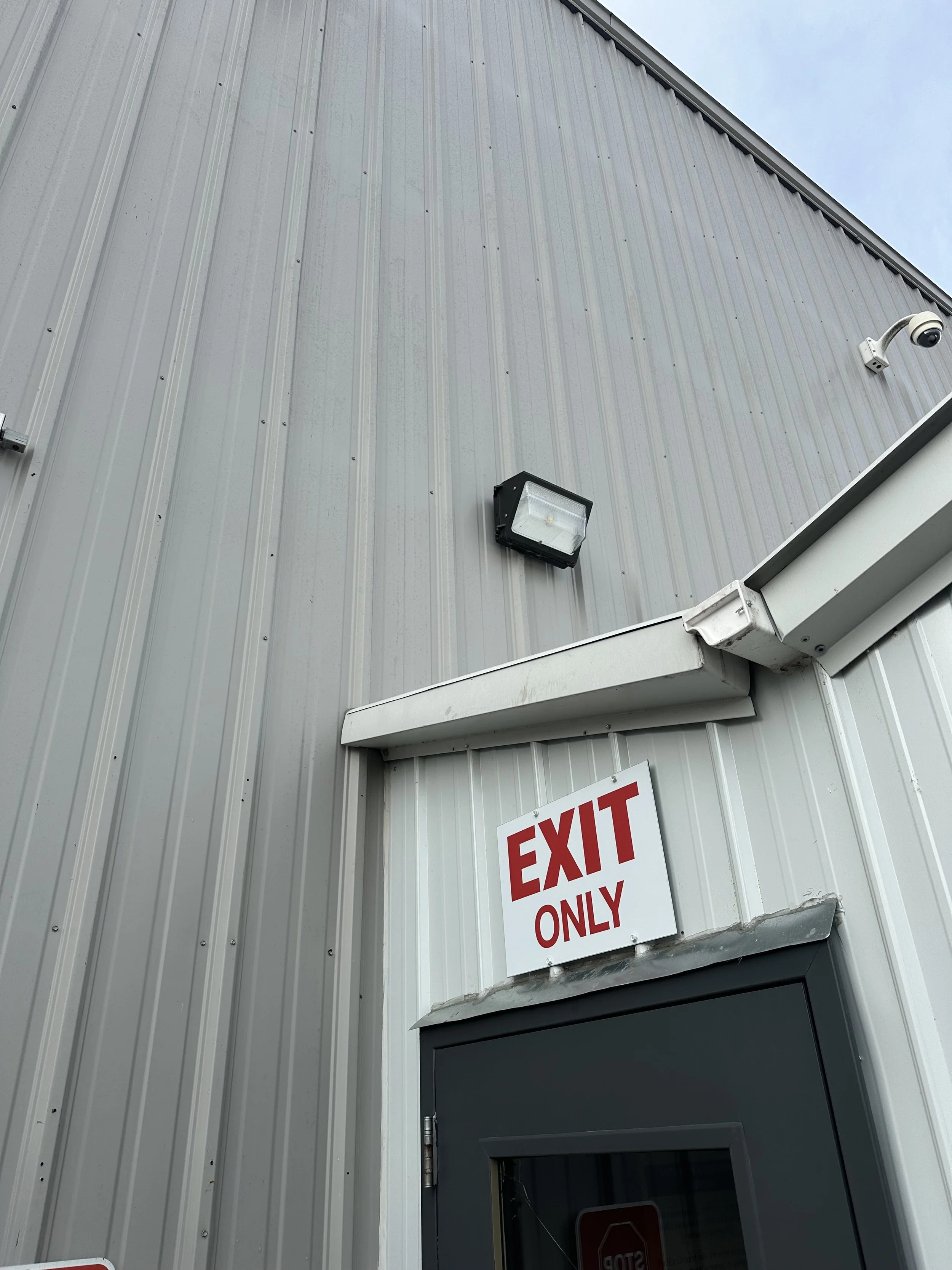Exterior of a building with a gray metal siding, security camera, floodlight, and a white sign with red letters reading 'EXIT ONLY'.