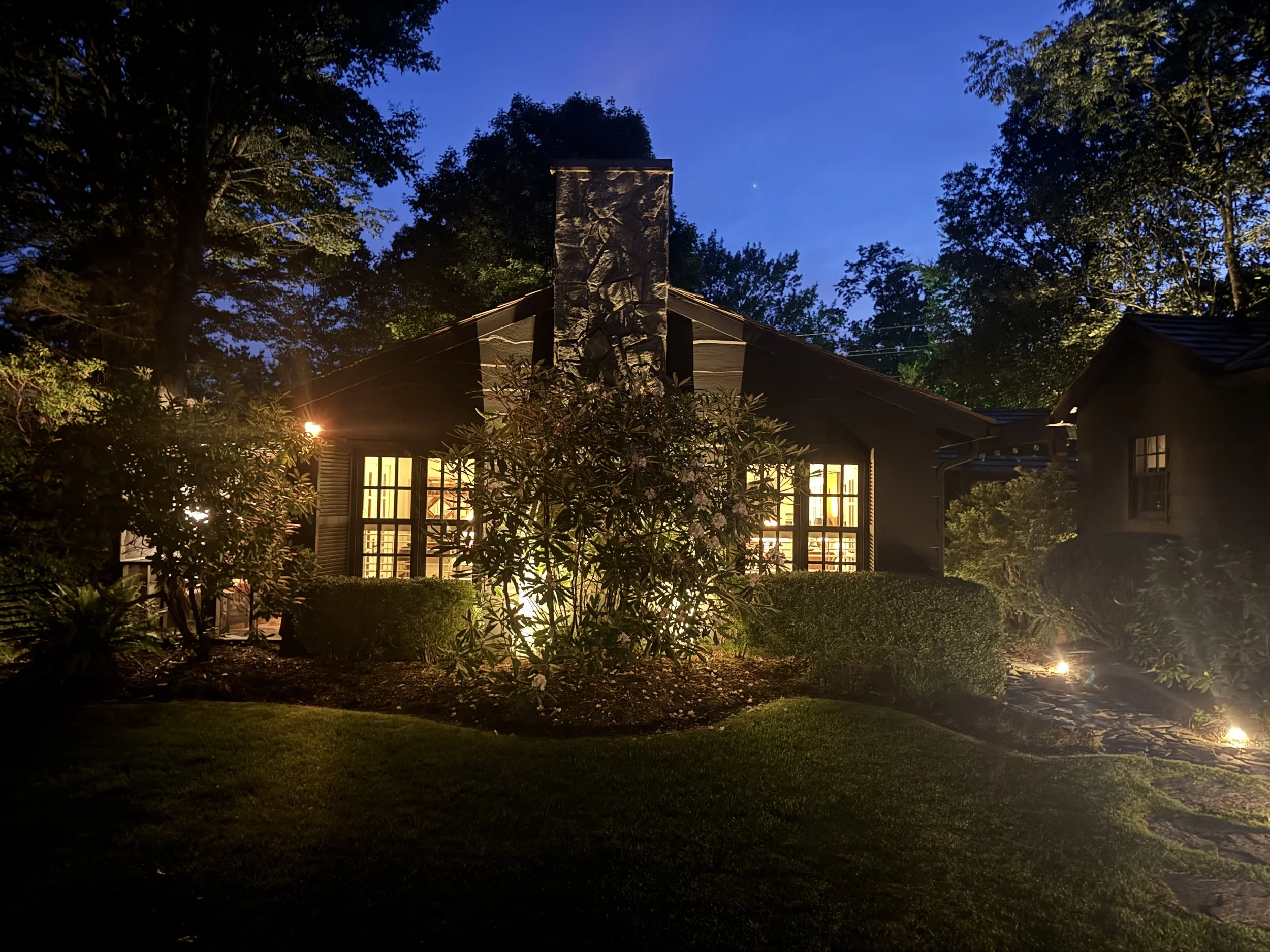 Nighttime view of a house with illuminated windows surrounded by trees and shrubs, with a dark blue sky and a bright star or planet visible.