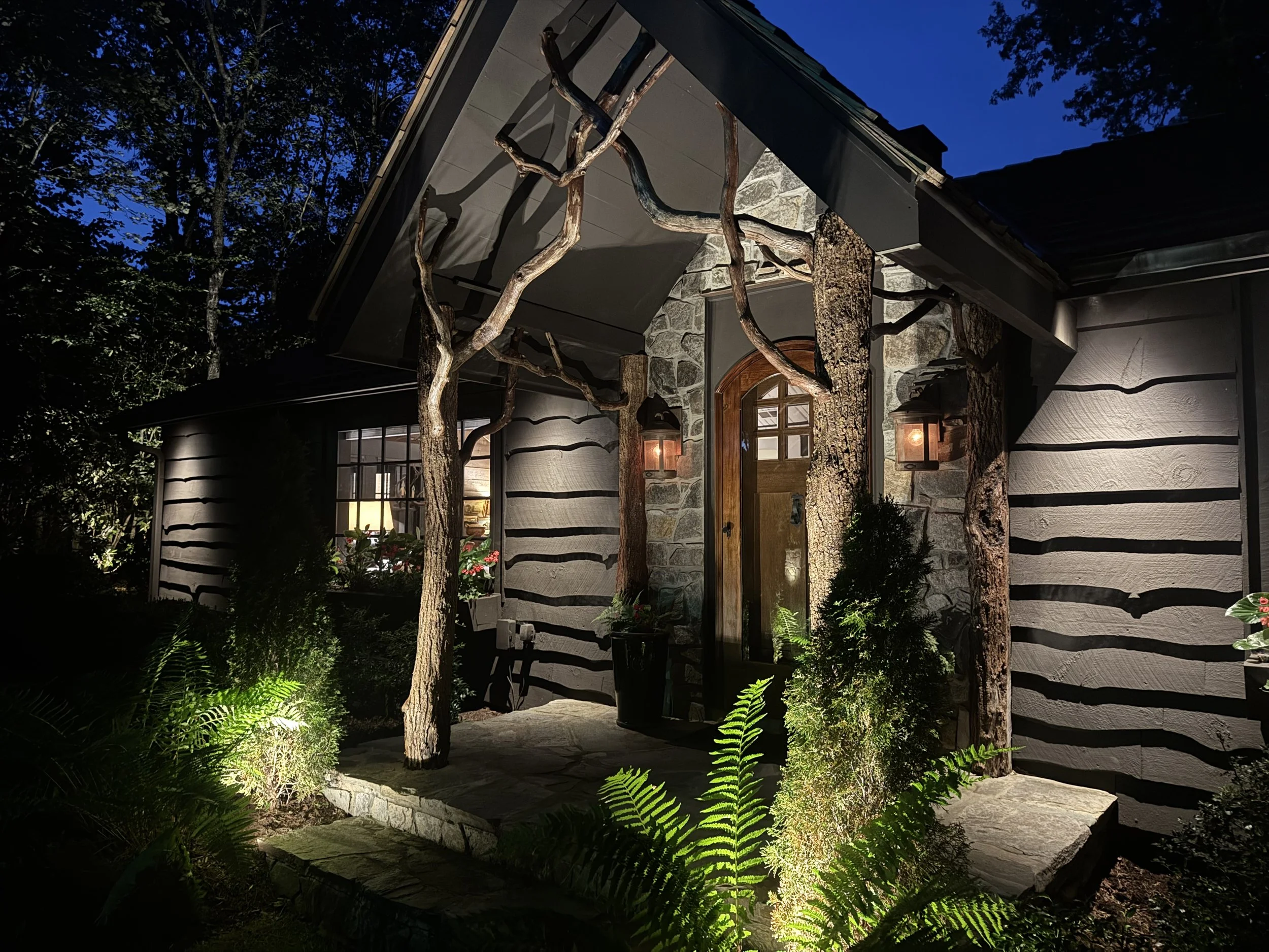 Nighttime view of a house with a stone and wood exterior, lit by outdoor lanterns, surrounded by green plants and trees, with a dark blue sky overhead.